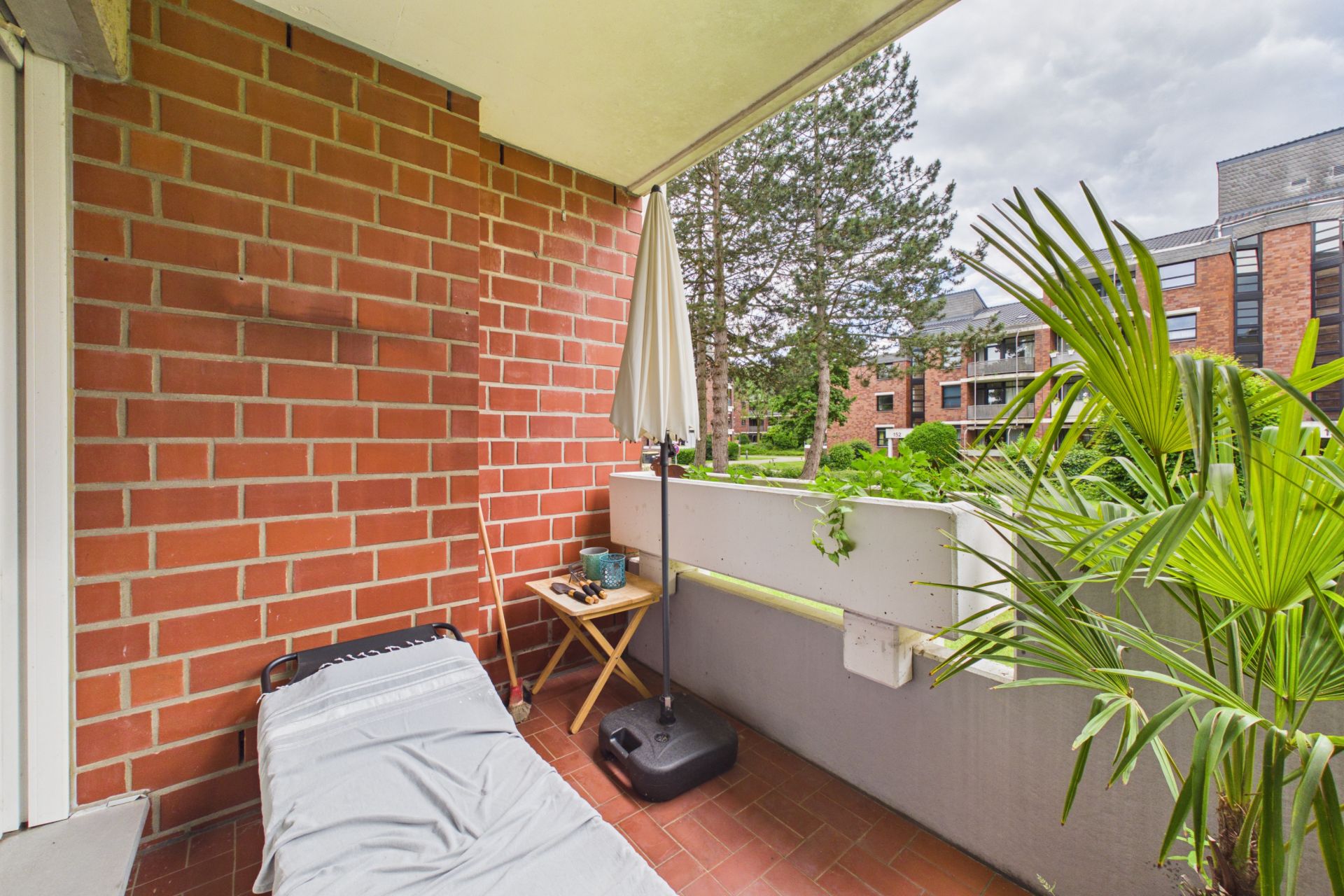Balkon mit Liege, kleinem Tisch, Sonnenschirm und Pflanze, Blick auf Bäume und Wohngebäude.