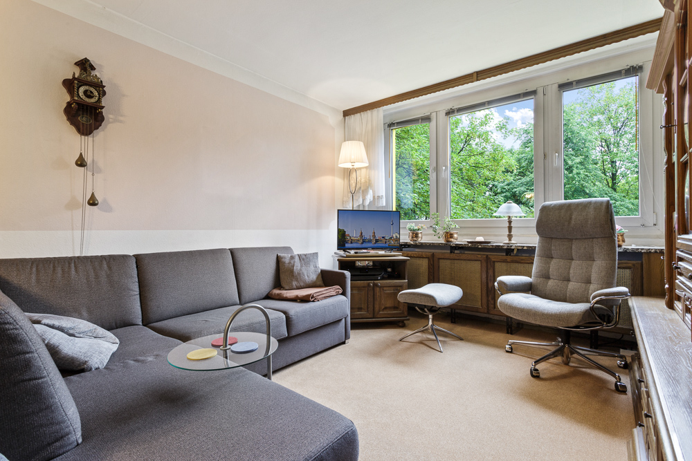 Wohnzimmer mit grauem Ecksofa, Sessel, Schreibtischstuhl und großen Fenstern mit Blick ins Grüne.