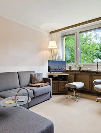 Wohnzimmer mit grauem Ecksofa, Sessel, Schreibtischstuhl und großen Fenstern mit Blick ins Grüne.