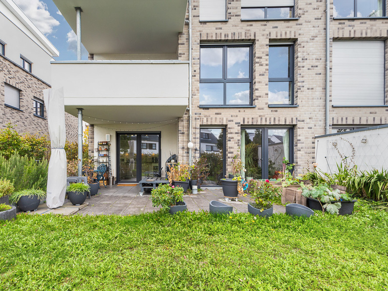 Moderne Hausfassade mit Terrasse, vielen Fenstern und Blumentöpfen im Garten.