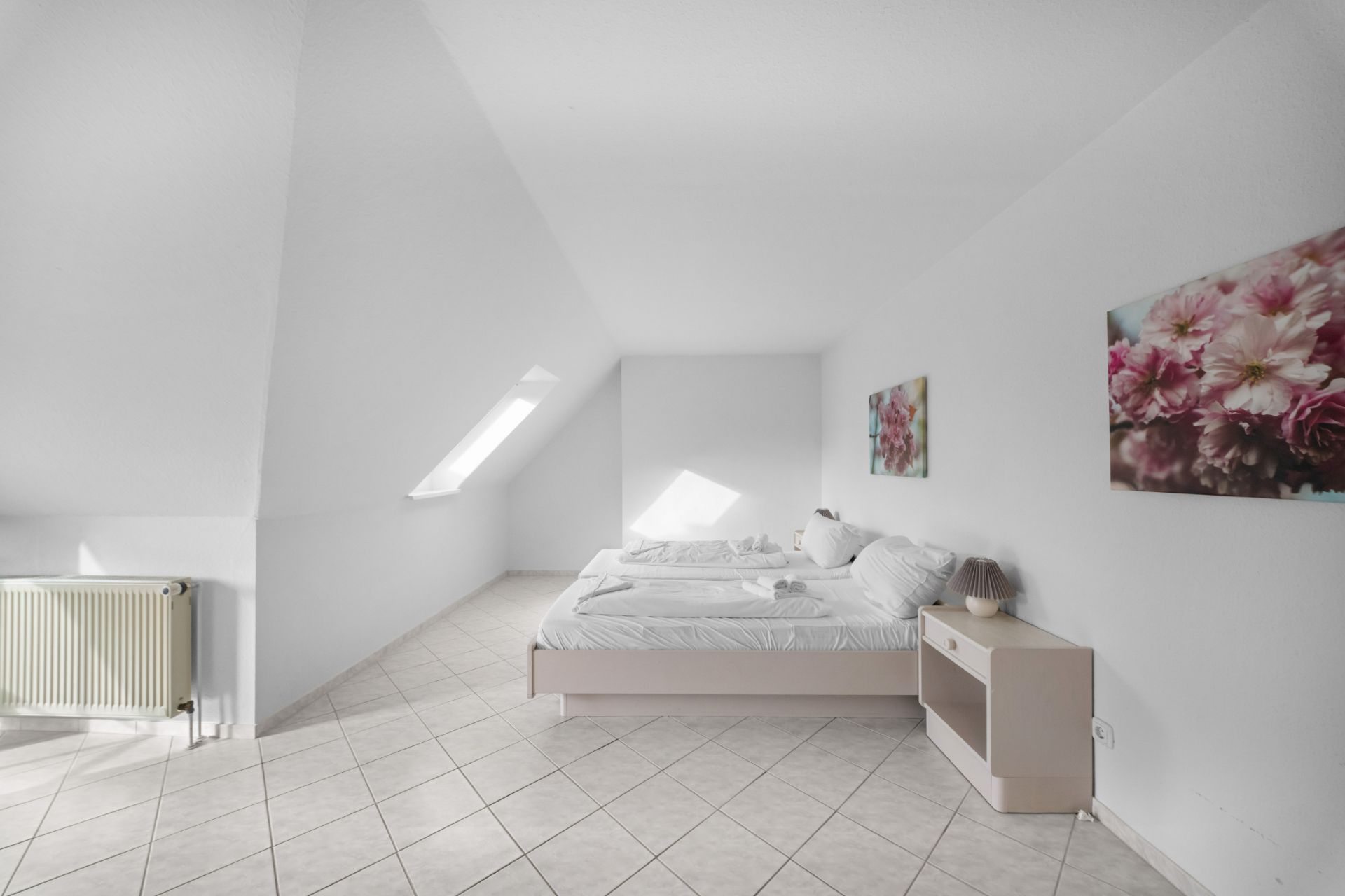 Helles Schlafzimmer mit Doppelbett, Nachttisch, Fliesenboden und Blumenbildern an der Wand.