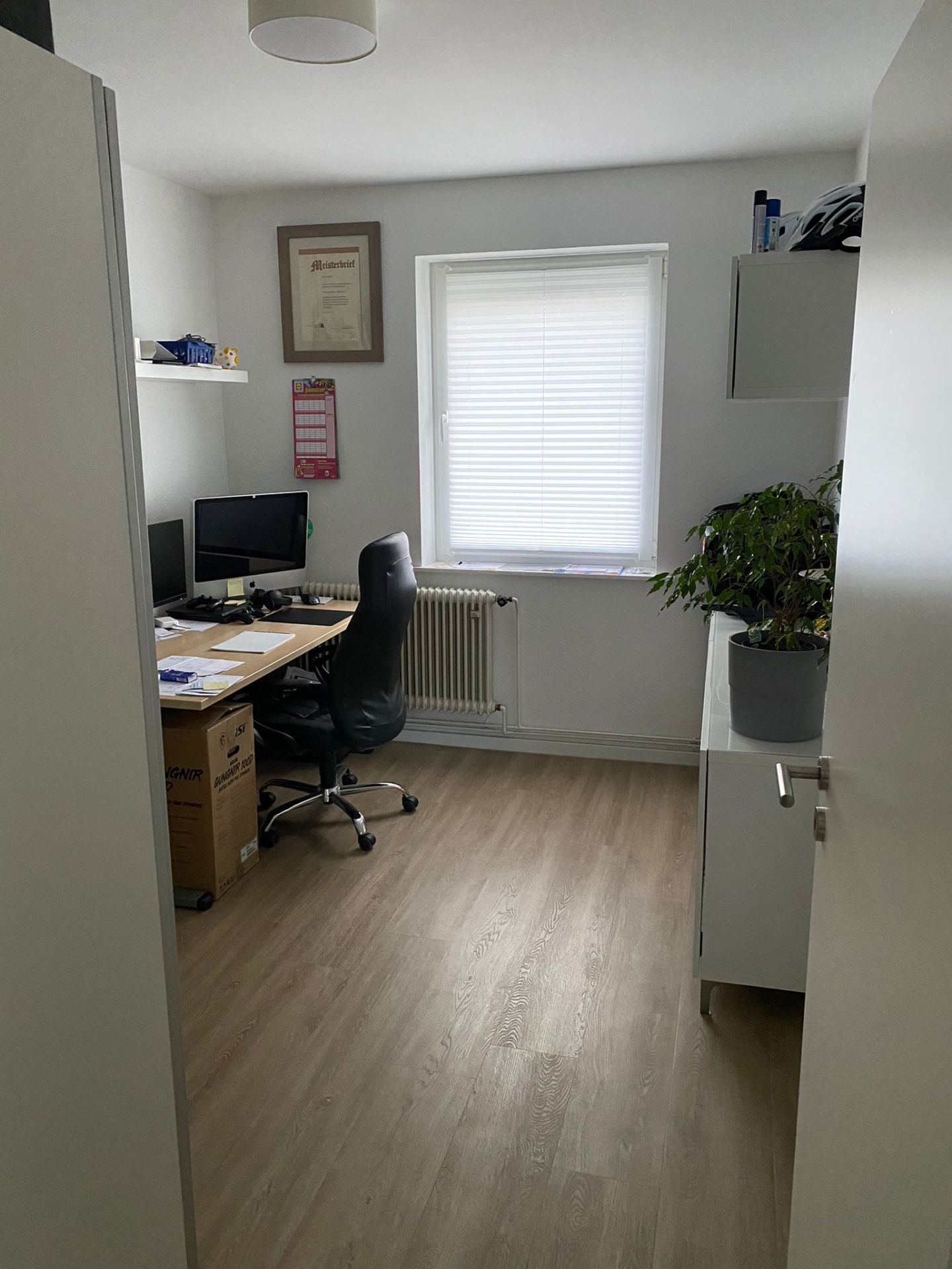 Arbeitszimmer mit Schreibtisch, Bürostuhl, Computer, Pflanze und Fenster mit Jalousien.