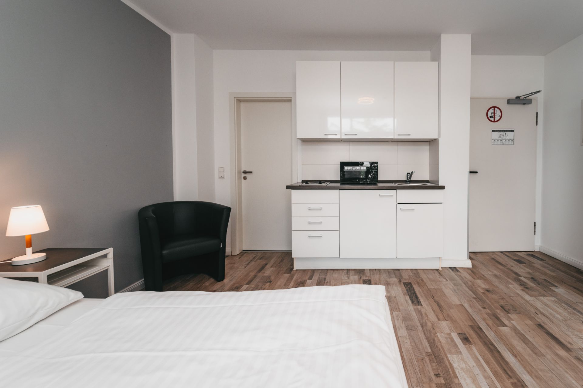 Helles Zimmer mit Bett, Nachttischlampe, Sessel und kleiner weißer Küchenzeile.