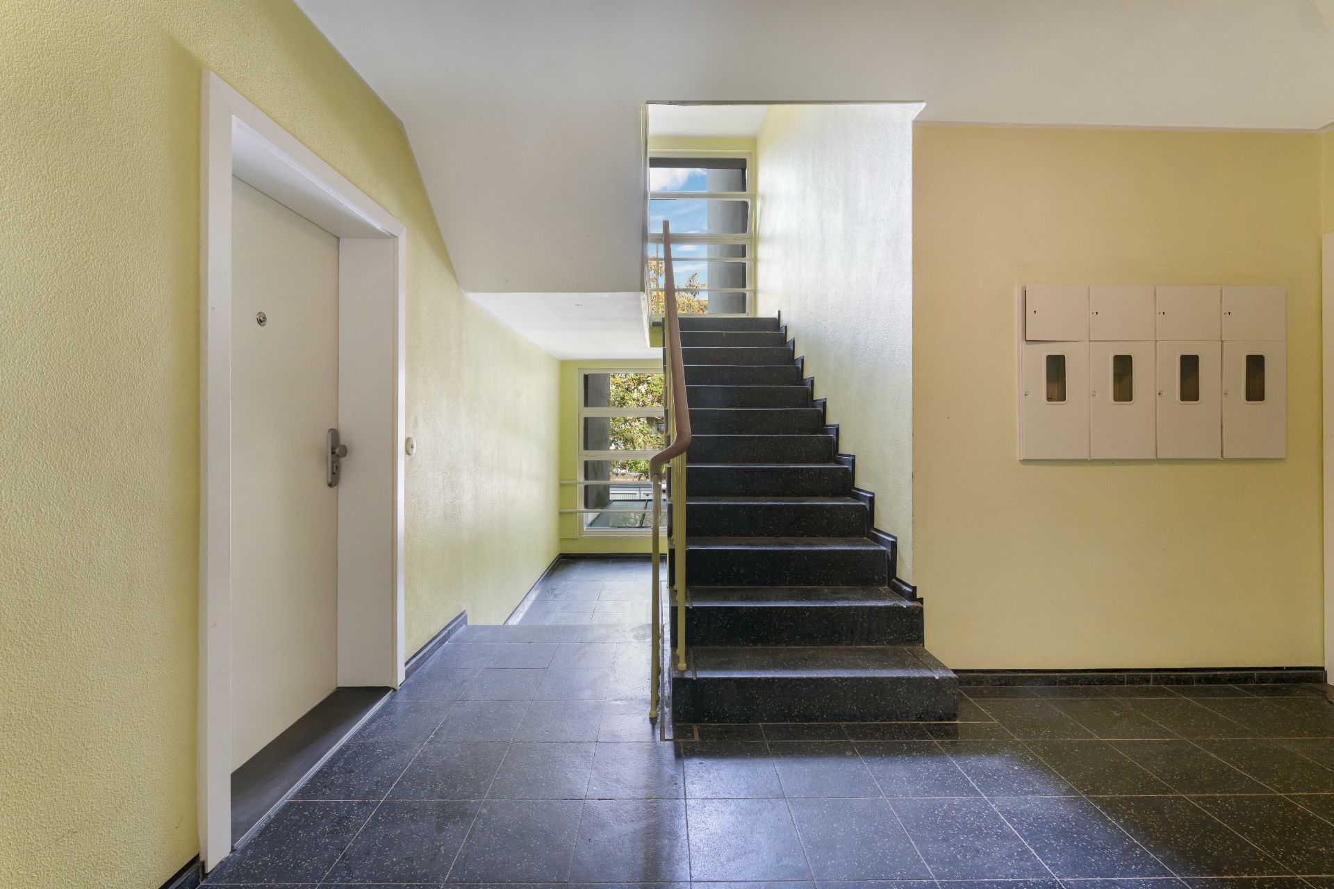 Hausflur mit Treppenaufgang, weißen Türen und Briefkästen an der gelben Wand.