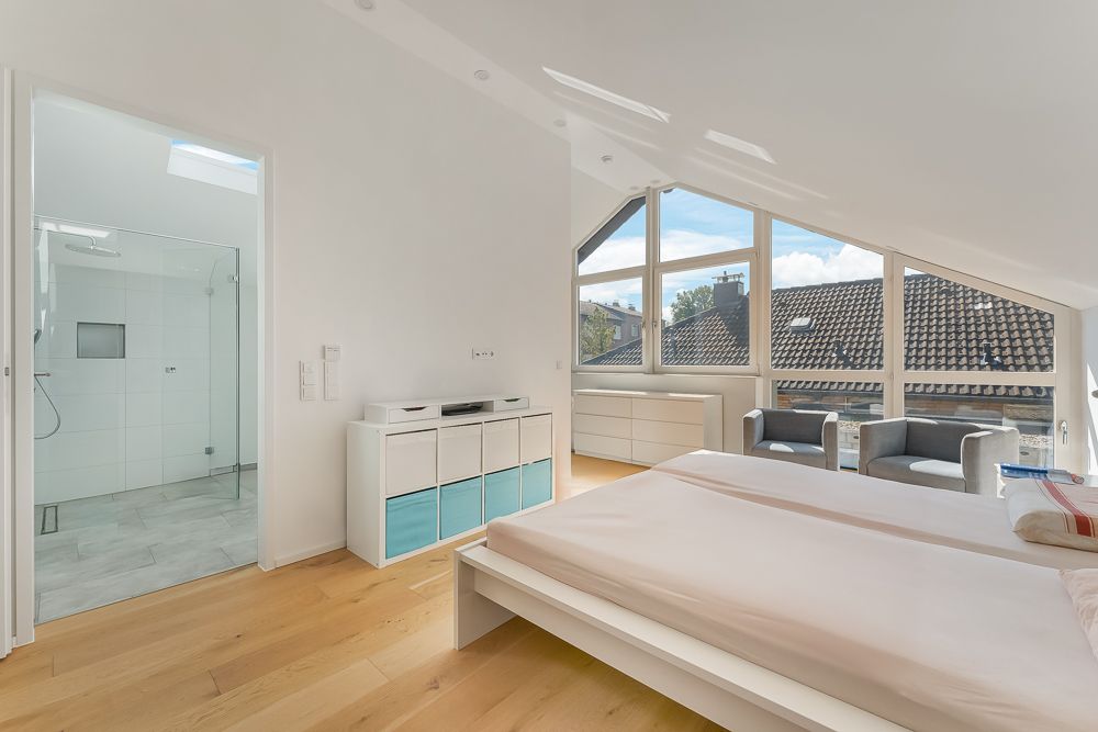 Helles Schlafzimmer mit großem Fenster, zwei Sesseln, Bett und Zugang zu einem Bad mit Dusche.