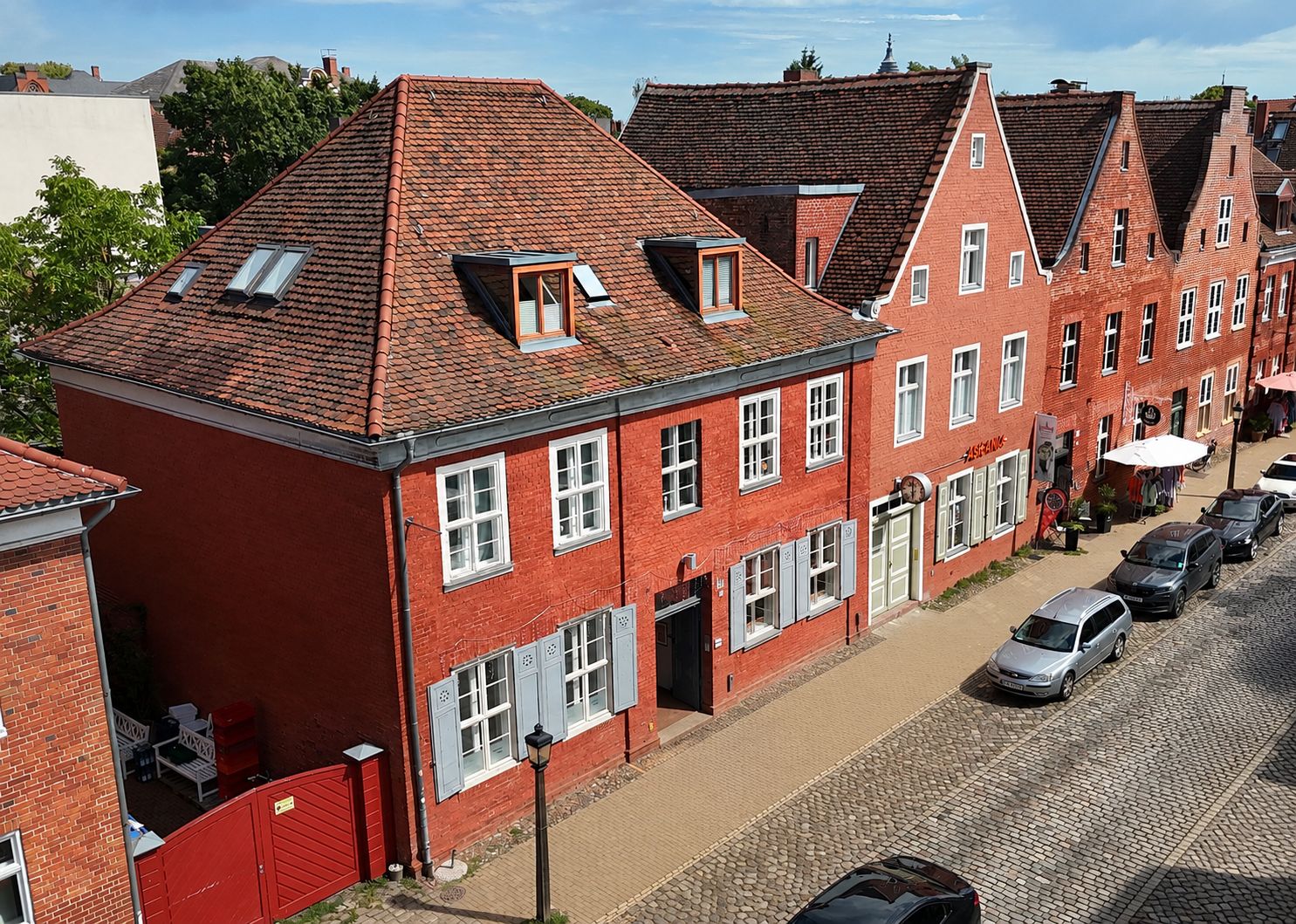 Rote Backsteinhäuser mit Satteldächern an einer Kopfsteinpflasterstraße, mehrere geparkte Autos.