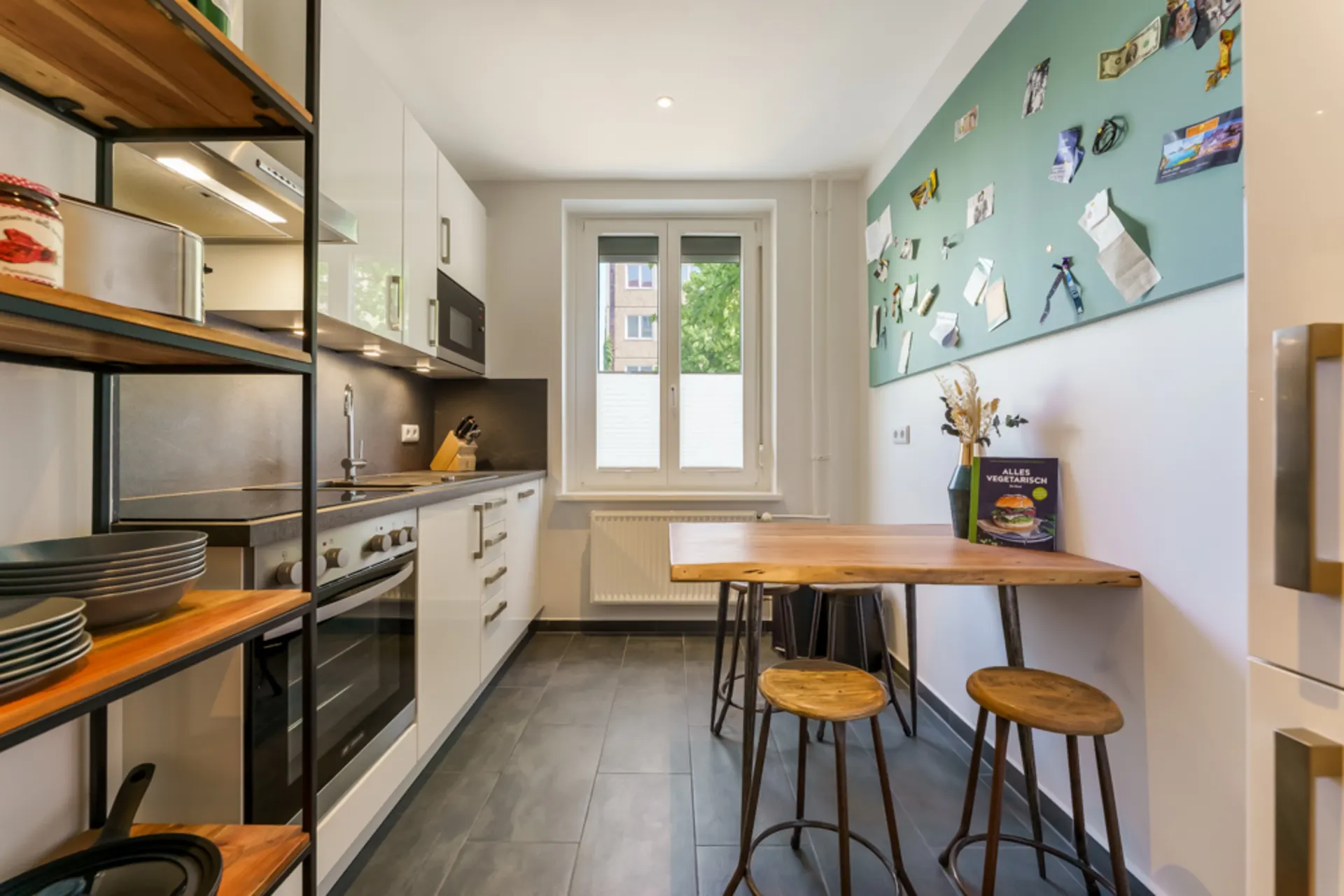 Moderne Küche mit Holztisch, drei Hockern, Wandtafel und großem Fenster.