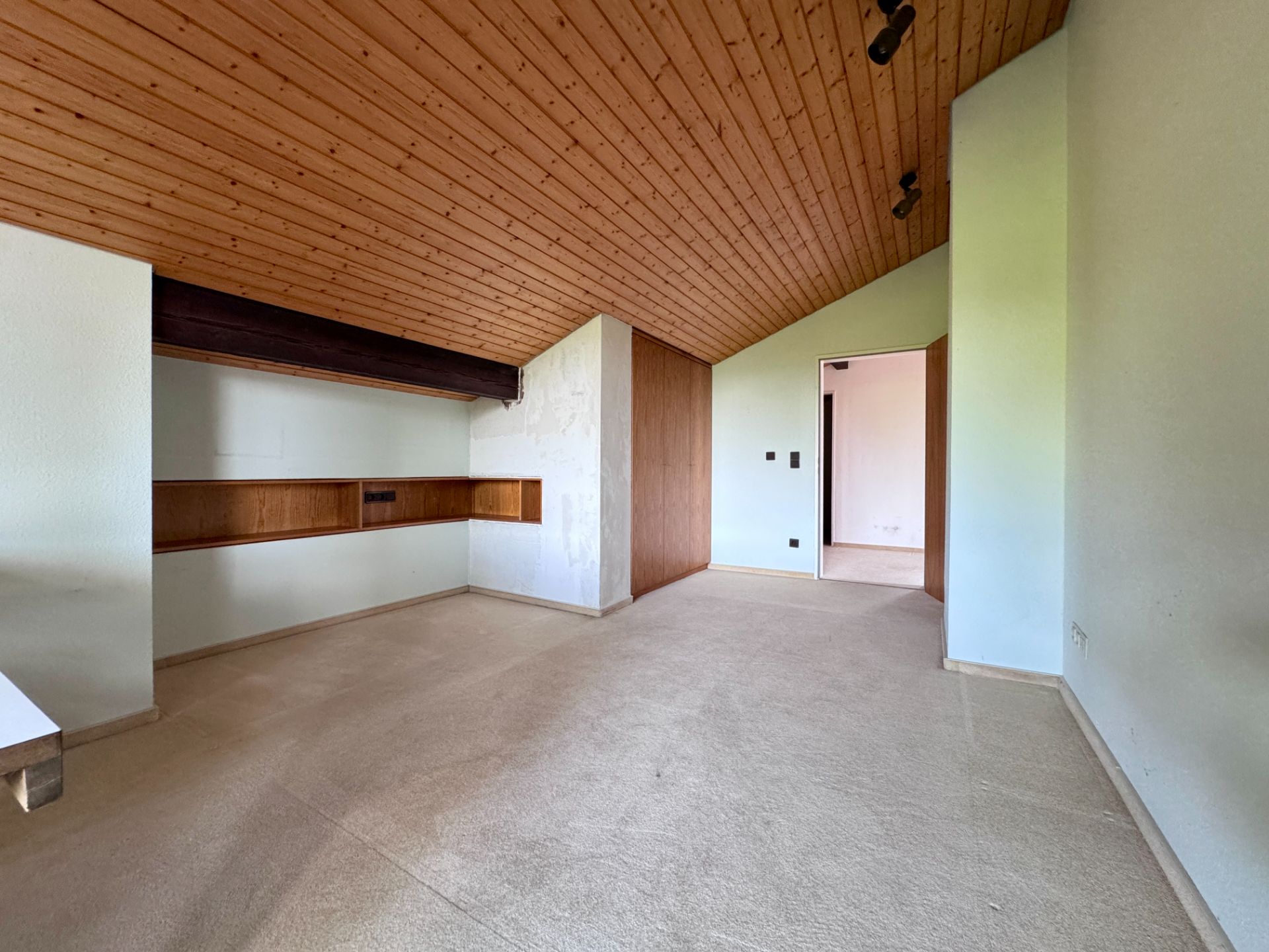 Leeres Zimmer mit schräger Holzdecke, beigem Teppich und Einbauschränken aus Holz.