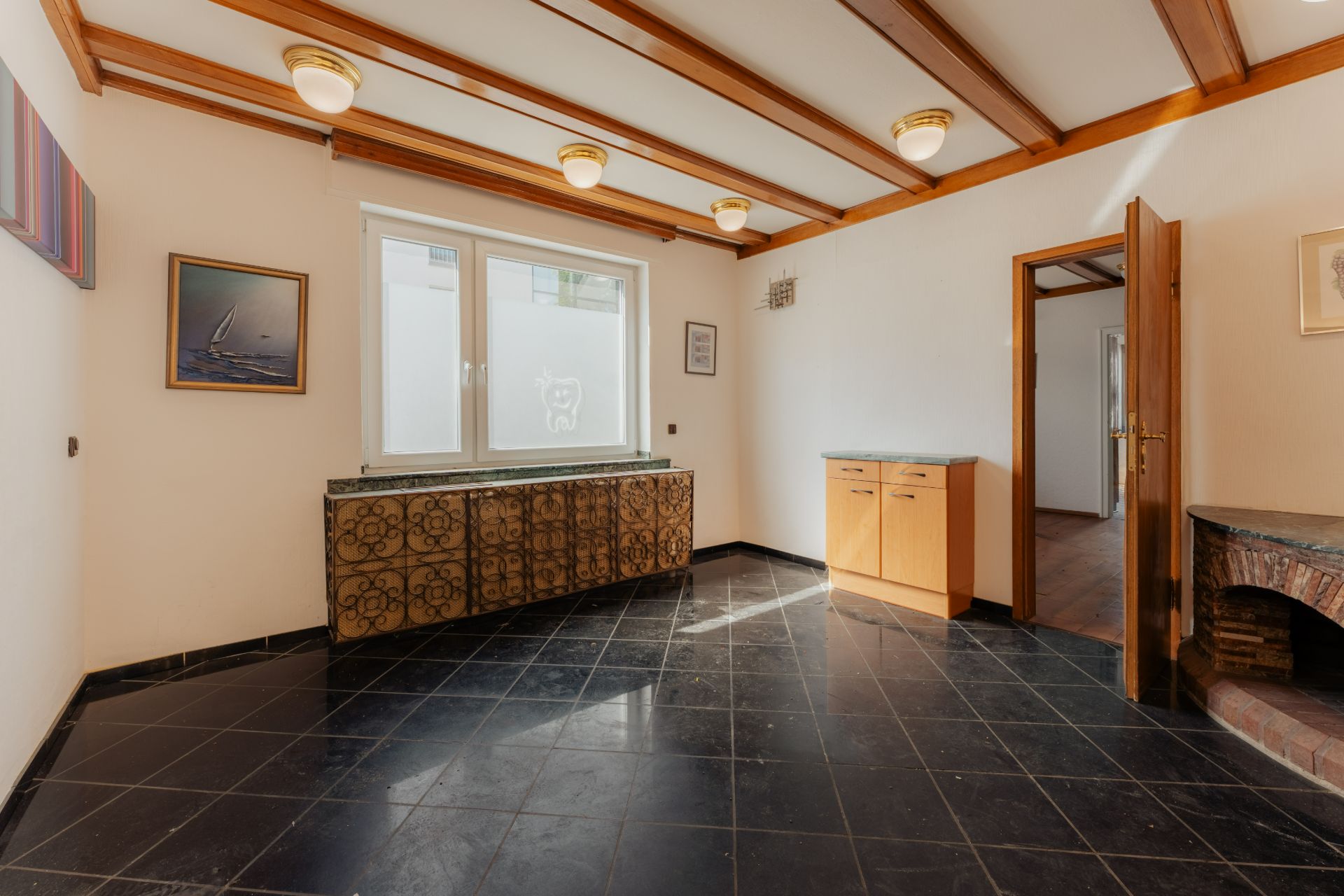 Heller Raum mit schwarzen Fliesen, Holzbalkendecke, Fenster, Bildern, Sideboard und offenem Durchgang.