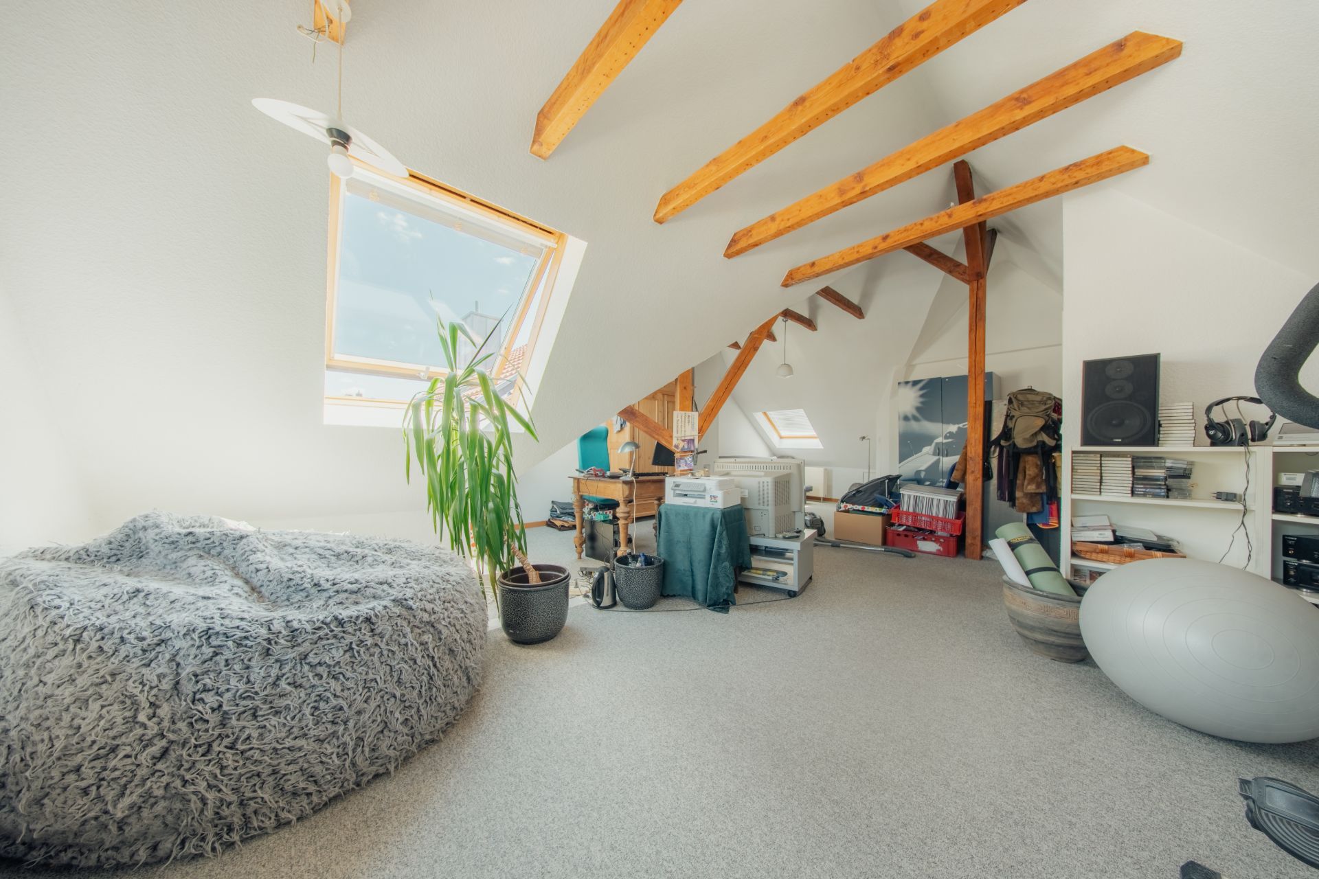 Helles Dachzimmer mit sichtbaren Holzbalken, Schreibtisch, Sitzsack, Pflanze und Regalen.