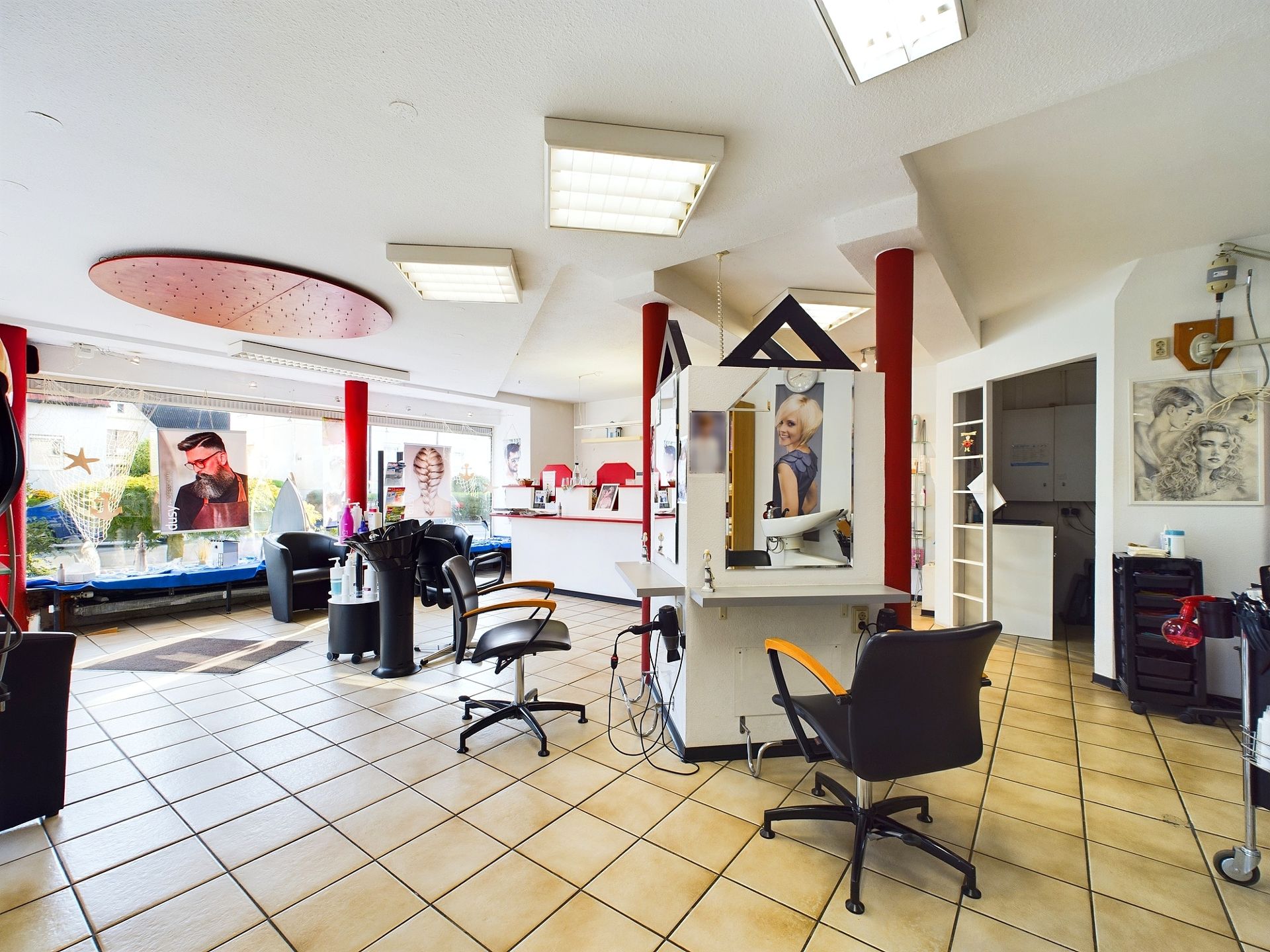 Heller Raum mit Friseurstühlen, Spiegeln und roten Säulen auf gefliestem Boden.