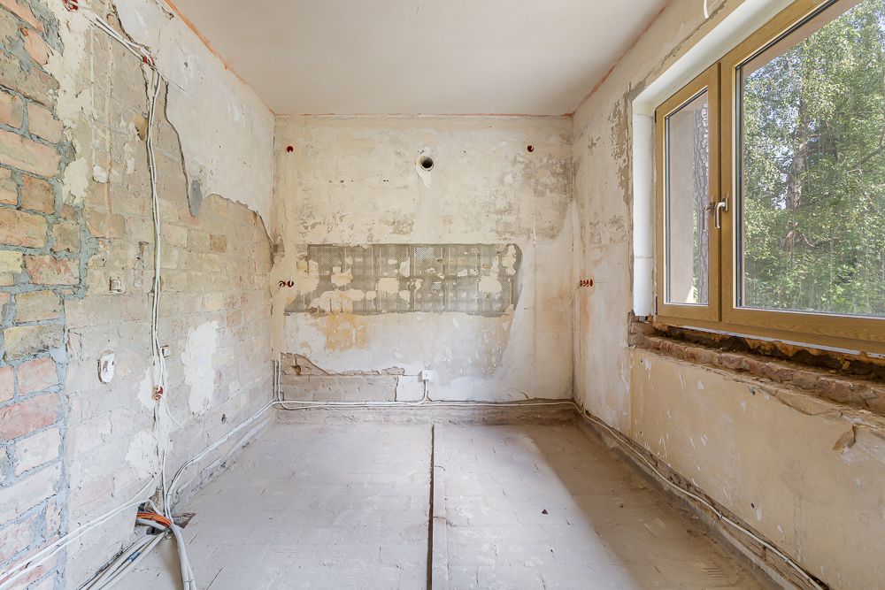Leerer, renovierungsbedürftiger Raum mit freigelegtem Mauerwerk und großem Fenster.