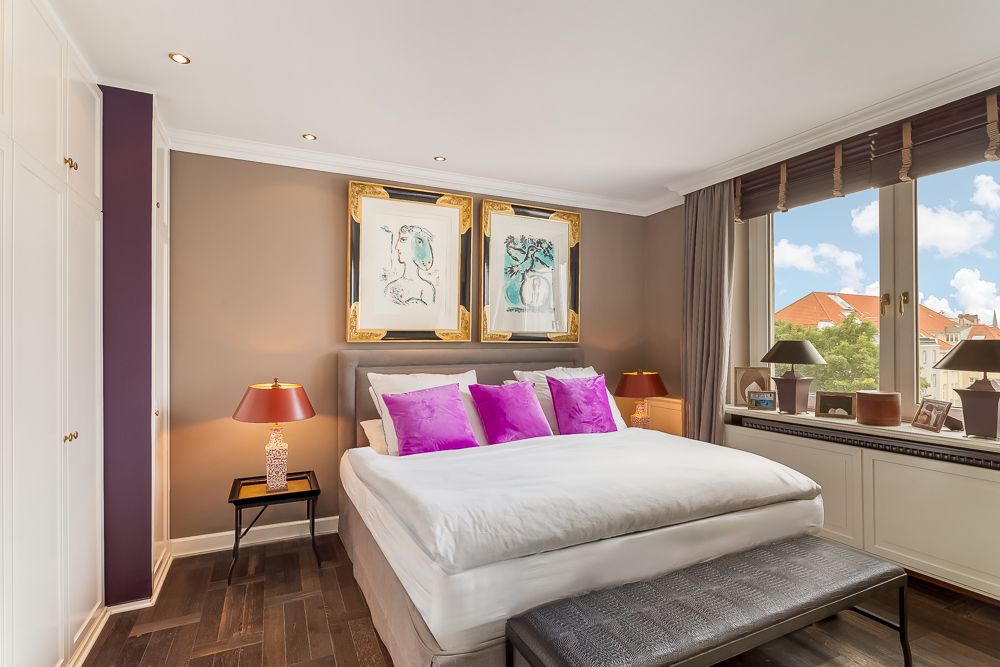 Helles Schlafzimmer mit Doppelbett, lila Kissen, großen Fenstern und zwei Bildern über dem Bett.