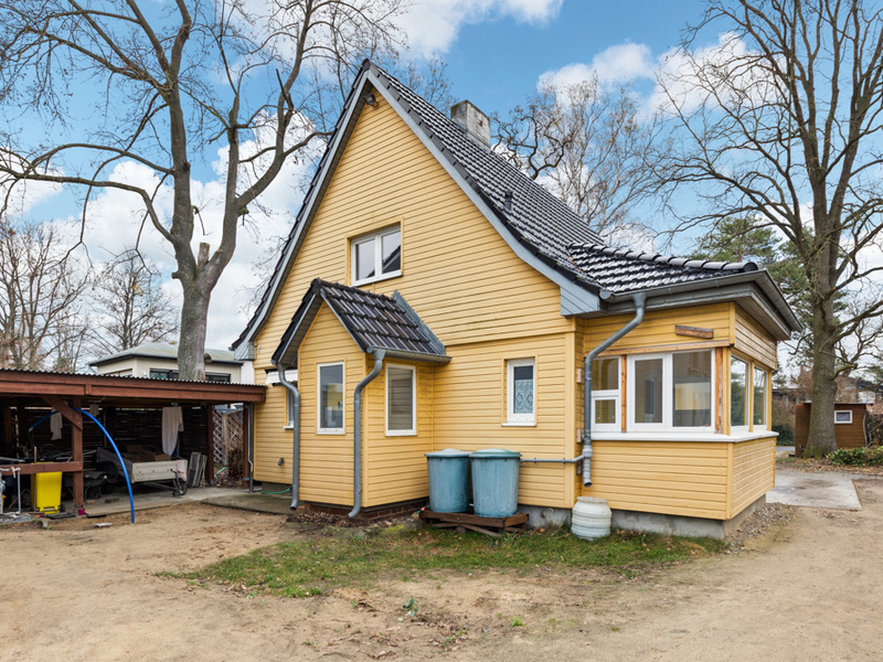 Gelbes Einfamilienhaus mit Satteldach, zwei Mülltonnen und Anbau, umgeben von Bäumen.