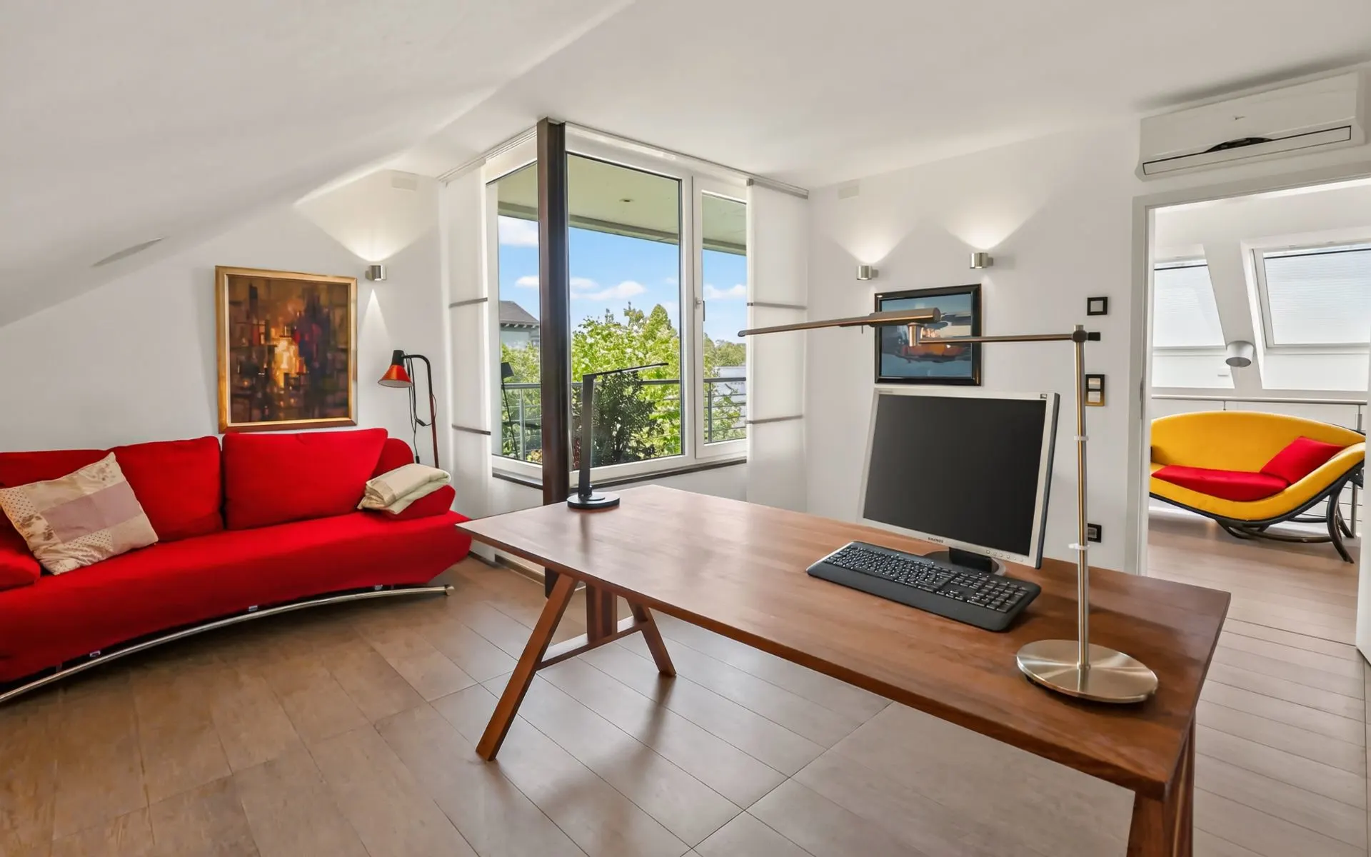 Helles Arbeitszimmer mit rotem Sofa, Holztisch, Computer und großem Fenster mit Blick ins Grüne.