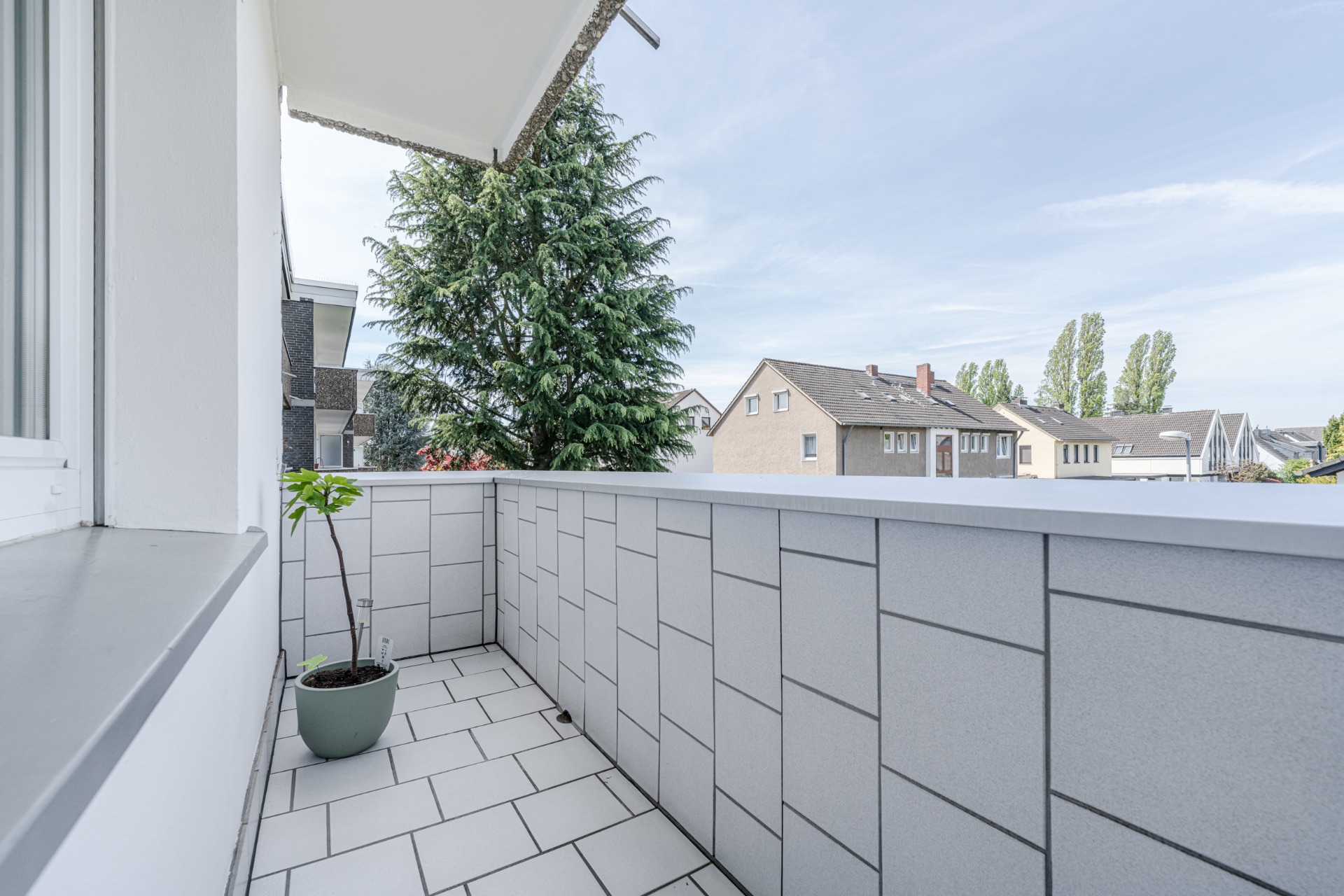Kleiner Balkon mit grauen Fliesen, einer Topfpflanze und Ausblick auf Wohnhäuser.