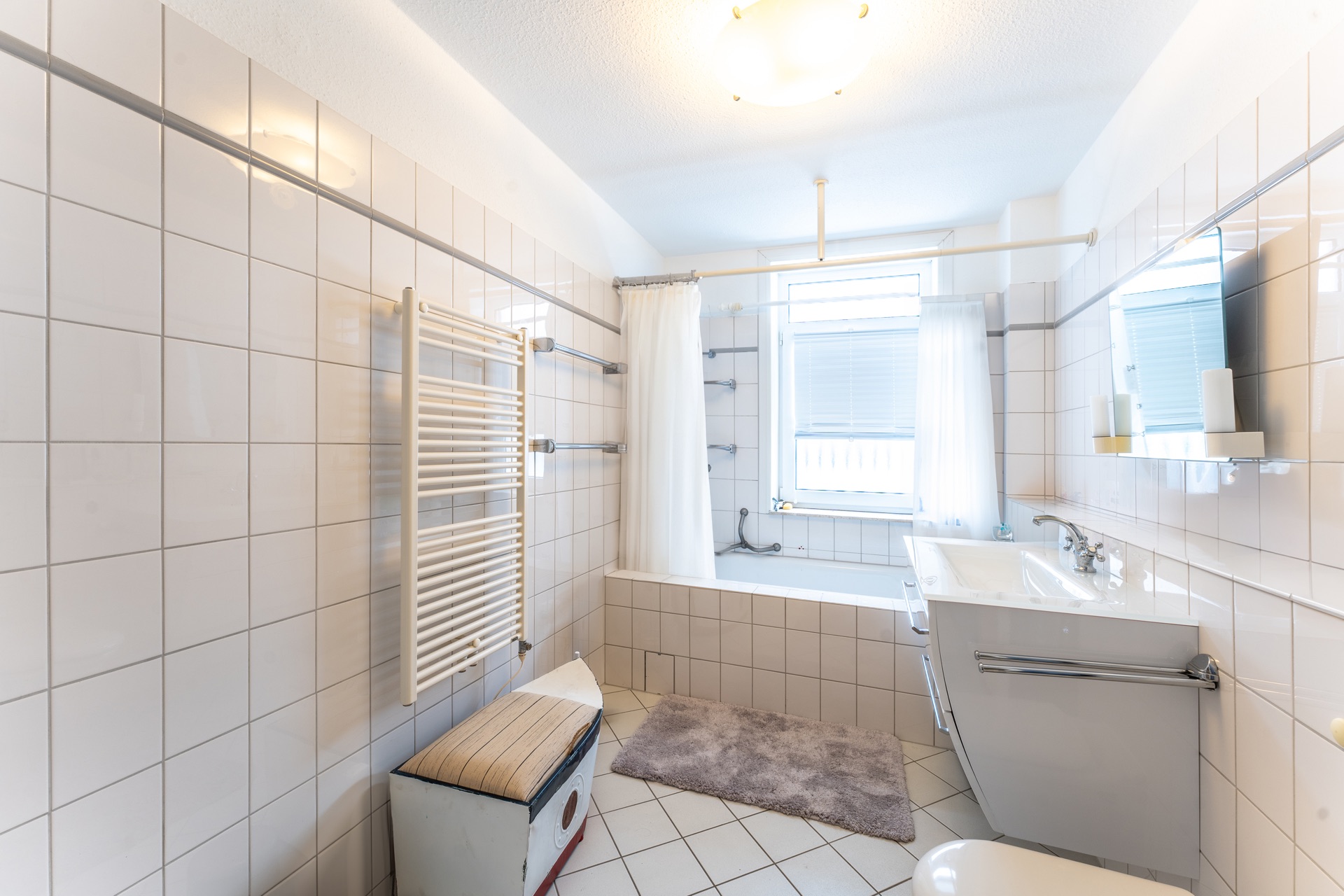 Helles Bad mit Badewanne, Waschbecken, Spiegel und Fenster, weiße Fliesen an Wand und Boden.