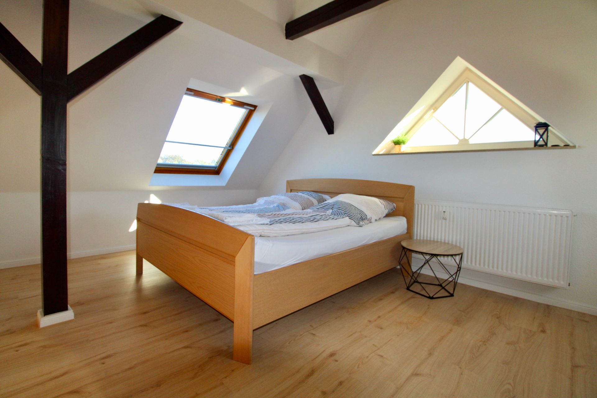 Dachzimmer mit Holzbett, schrägem Fenster, Heizung und kleinem runden Tisch auf hellem Holzboden.