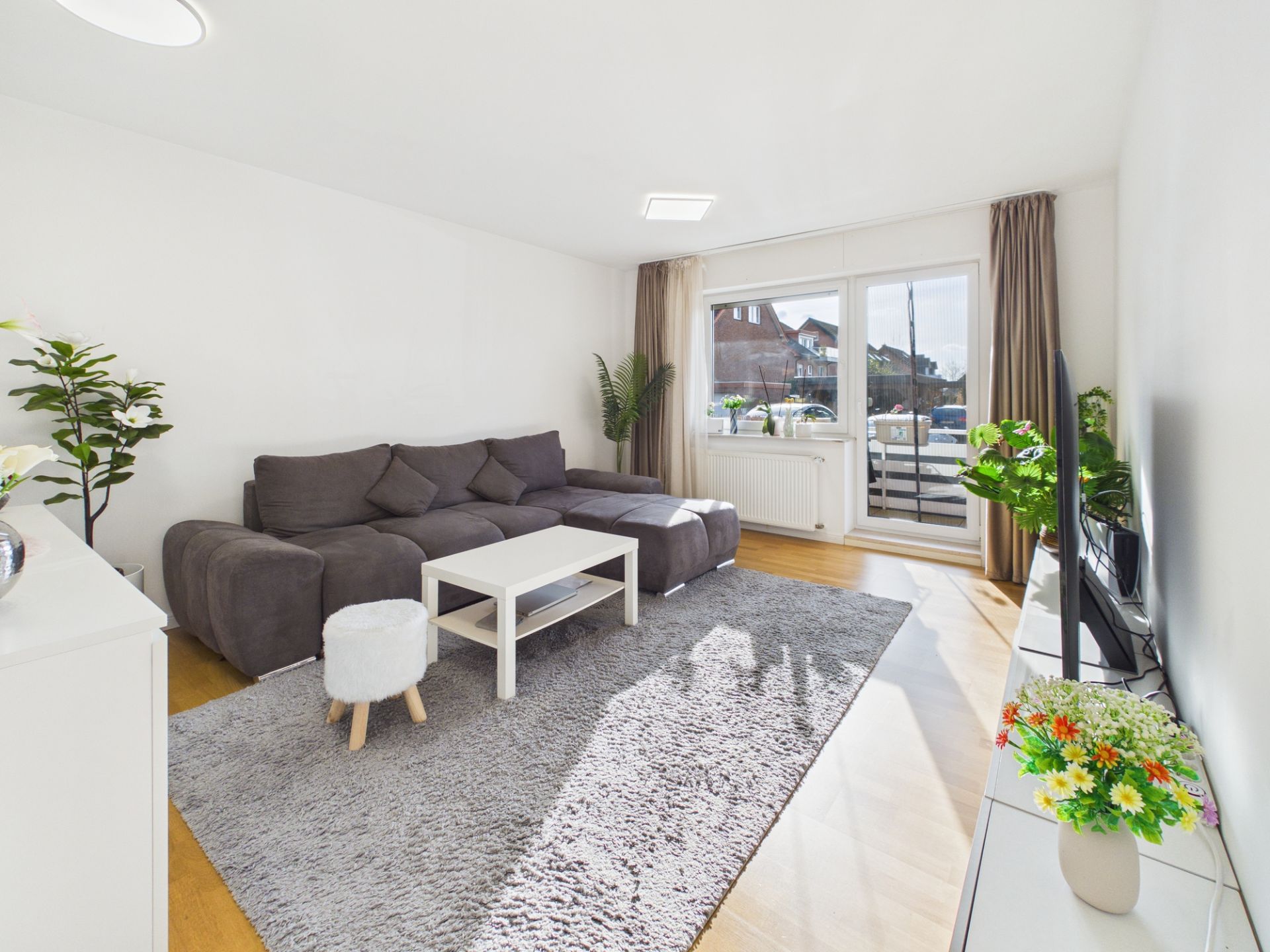 Helles Wohnzimmer mit grauem Sofa, Teppich, Couchtisch und Fernseher. Glas­tür führt auf einen Balkon.