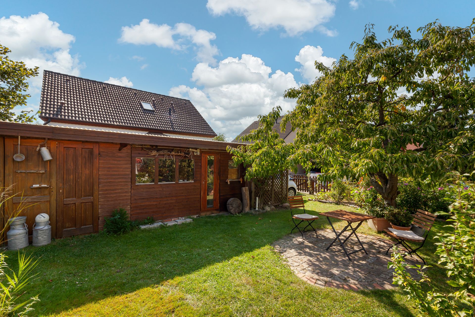 Holzhaus mit Garten, Baum und Sitzbereich mit Tisch und zwei Stühlen auf gepflastertem Untergrund.