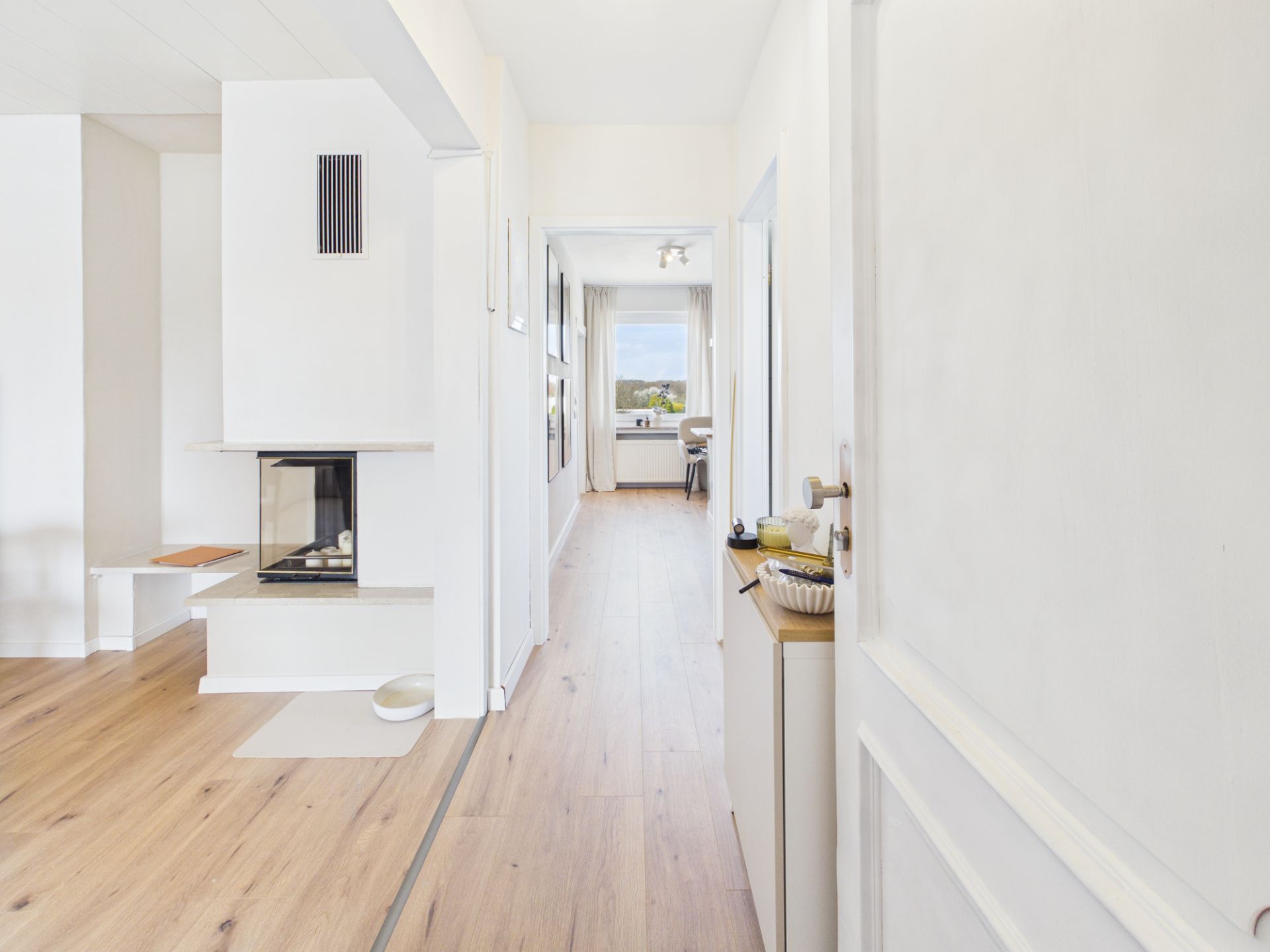 Heller Flur mit Holzboden, weißer Wand und Blick in ein Zimmer mit Fenster.