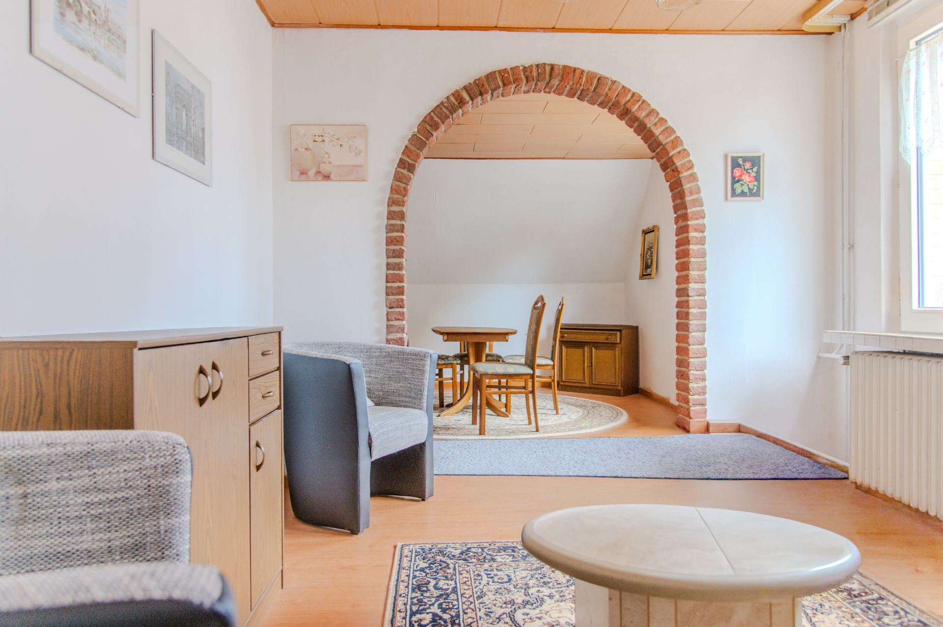 Wohnzimmer mit Sessel, Kommode und Rundbogen zu Essbereich mit Tisch und Stühlen.