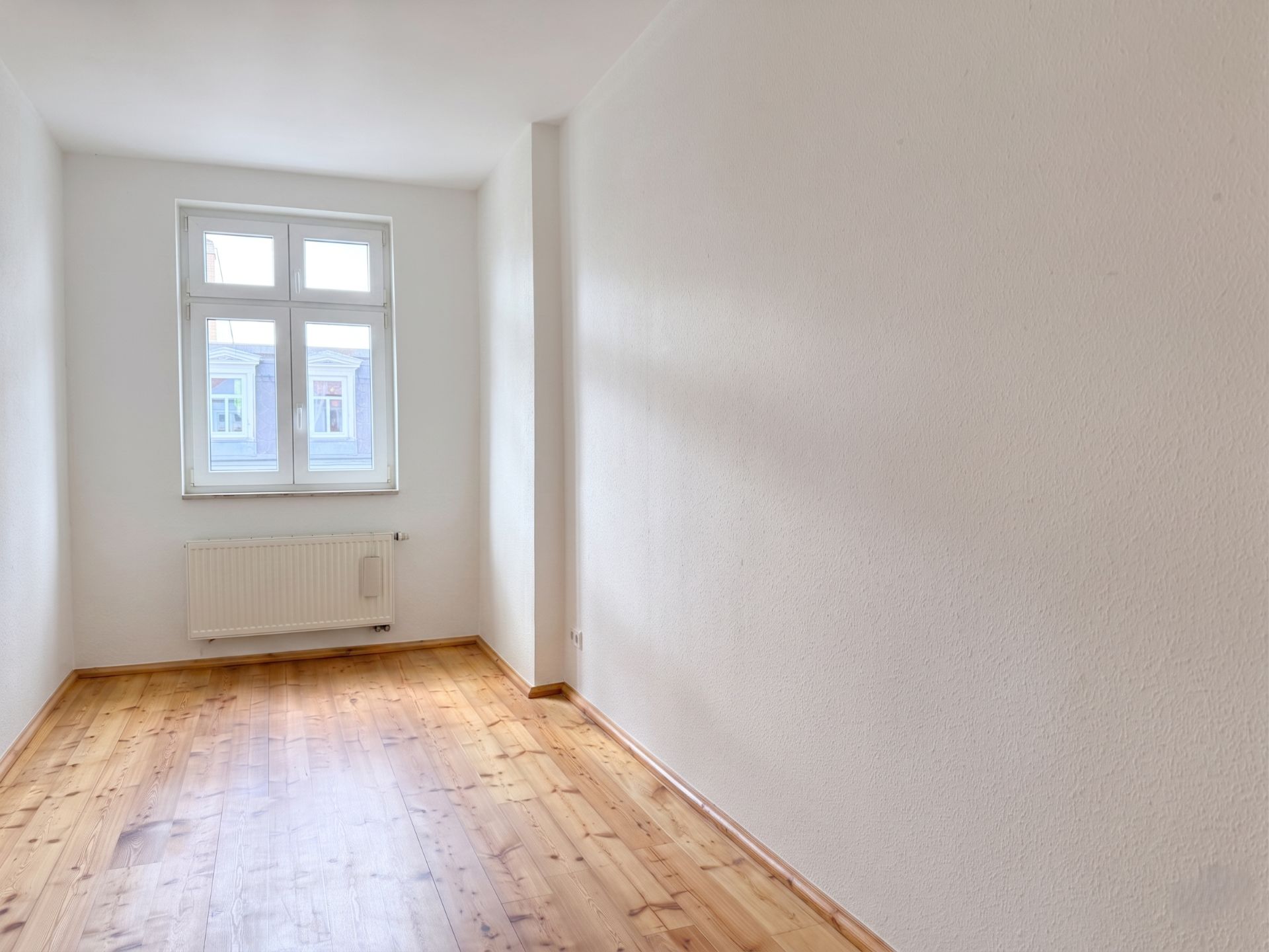 Schmaler, leerer Raum mit Holzboden, weißer Wand und Fenster mit Heizkörper.