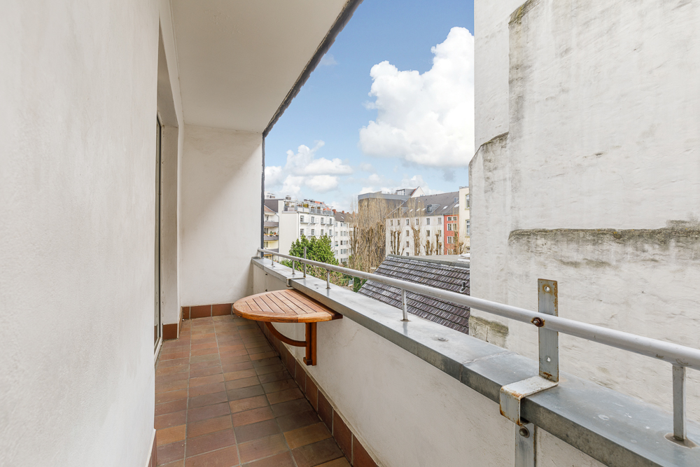 Balkon mit kleinem Holztisch, Fliesenboden und Blick auf angrenzende Gebäude.