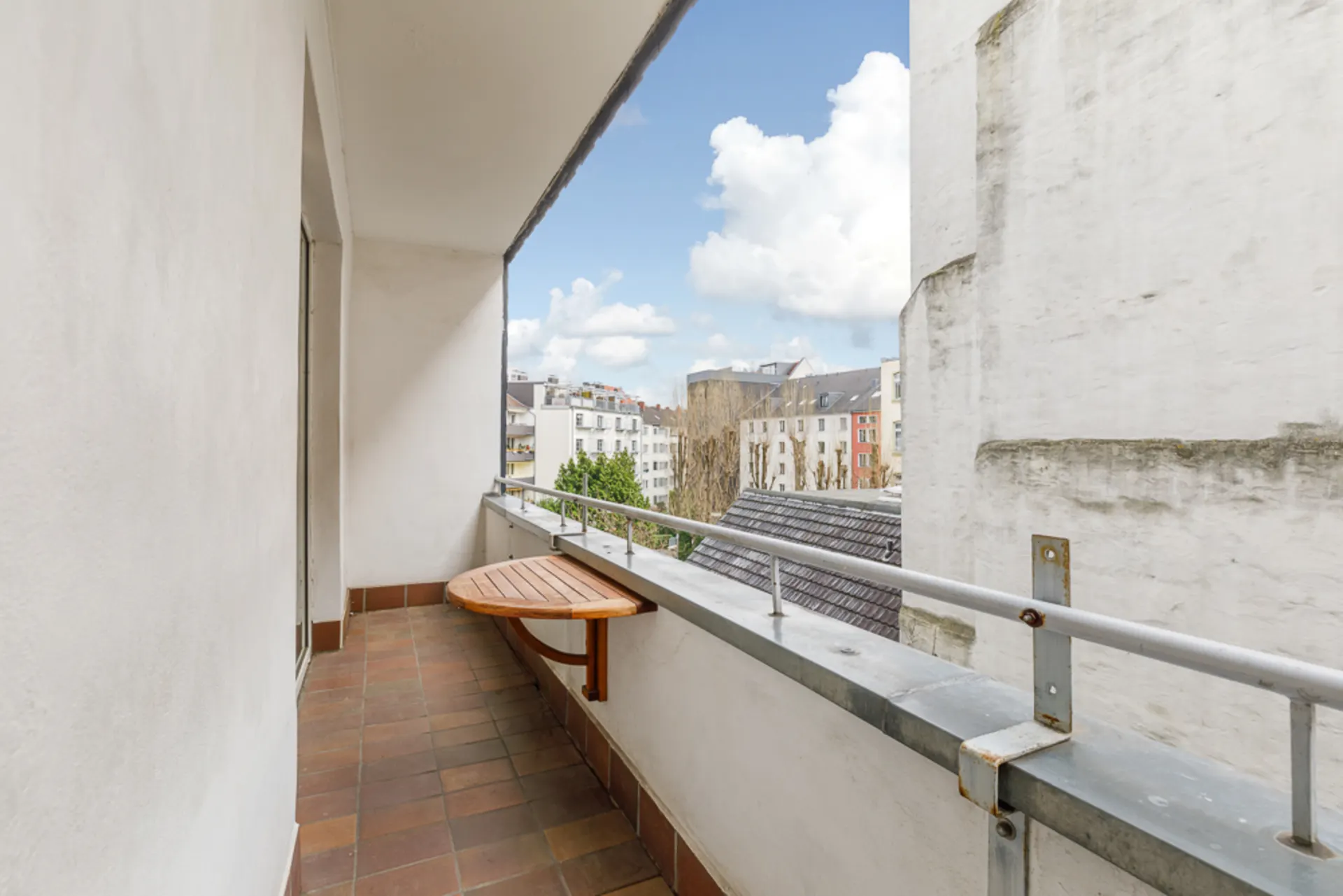 Balkon mit kleinem Holztisch, Fliesenboden und Blick auf angrenzende Gebäude.