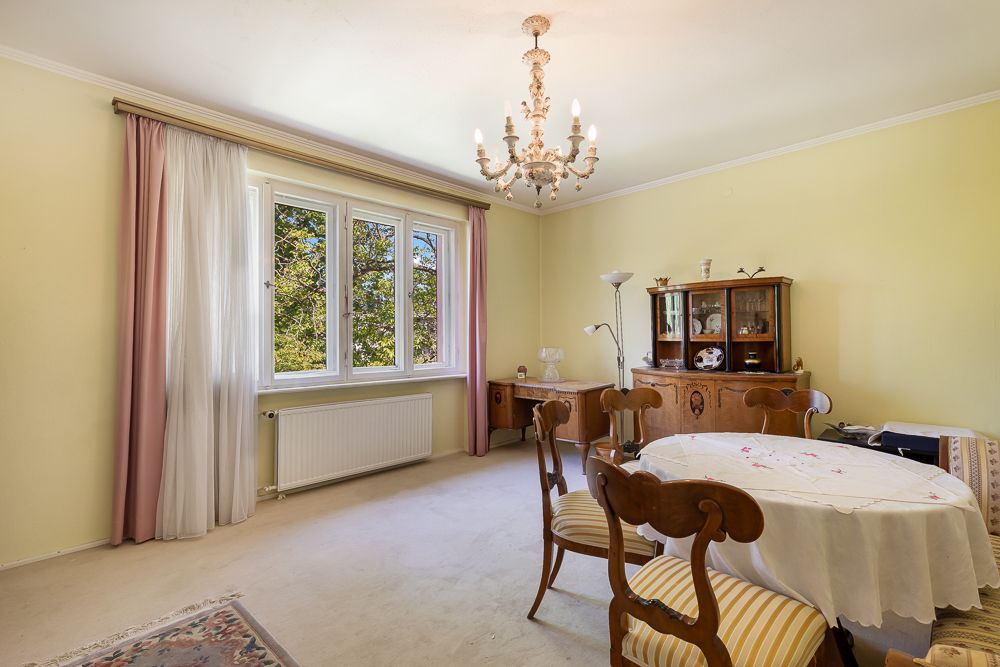 Wohnzimmer mit rundem Tisch, vier Stühlen, Kronleuchter und großem Fenster mit Vorhängen.