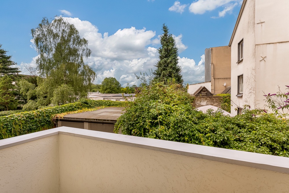 Blick von einem Balkon auf begrünte Dächer und Bäume bei sonnigem Wetter.