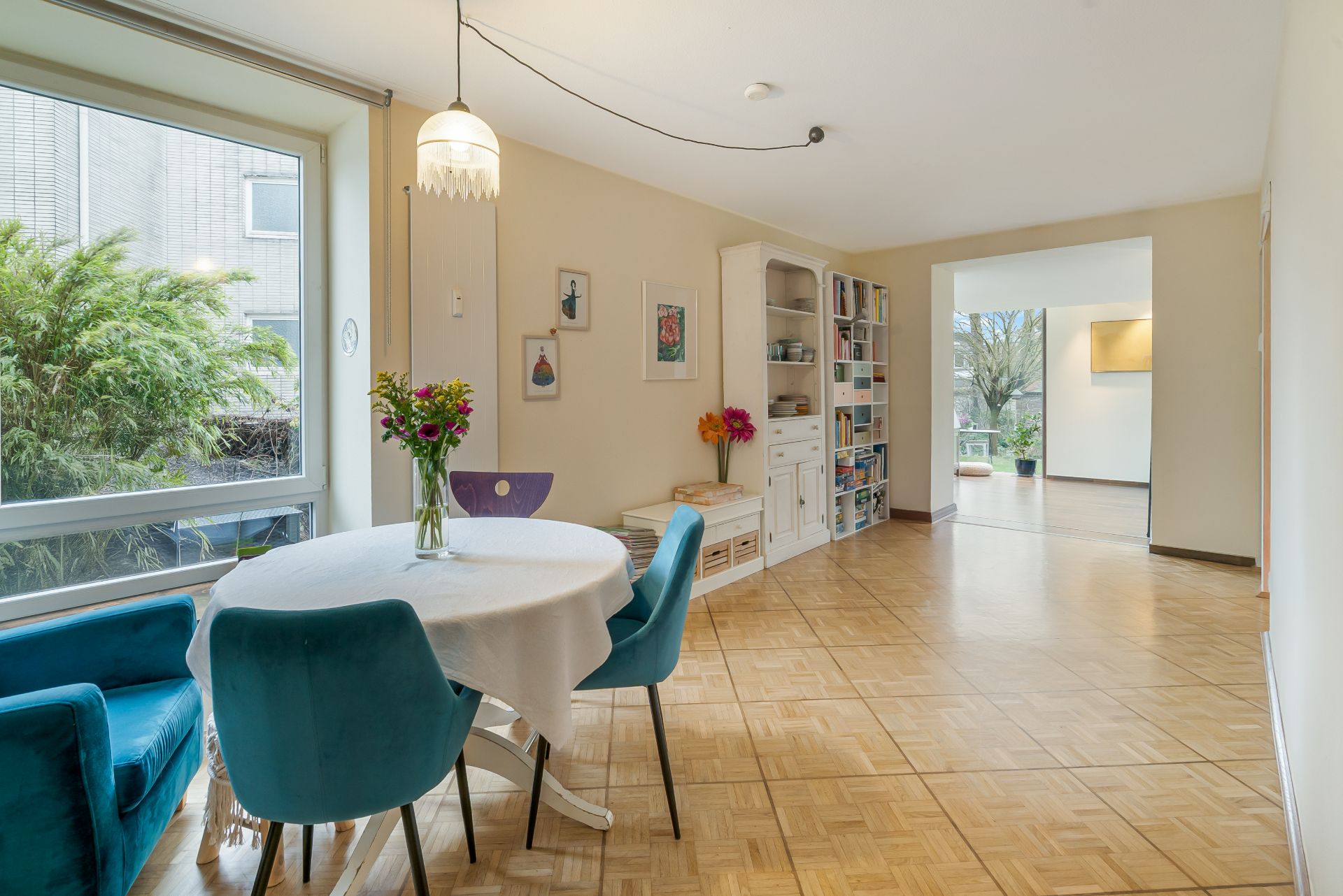 Essbereich mit rundem Tisch und blauen Stühlen, großes Fenster, Regal und Parkettboden.