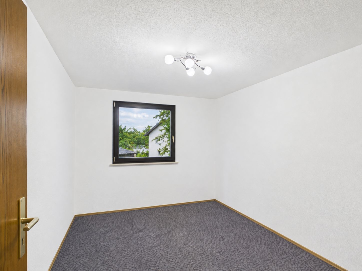 Leeres Zimmer mit weiß gestrichenen Wänden, Teppichboden und Fenster, durch das Bäume zu sehen sind.