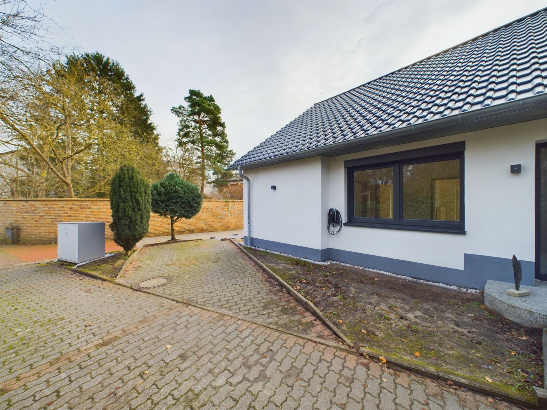 Gepflasterter Hof neben einem weißen Wohnhaus mit Garten und Bäumen.