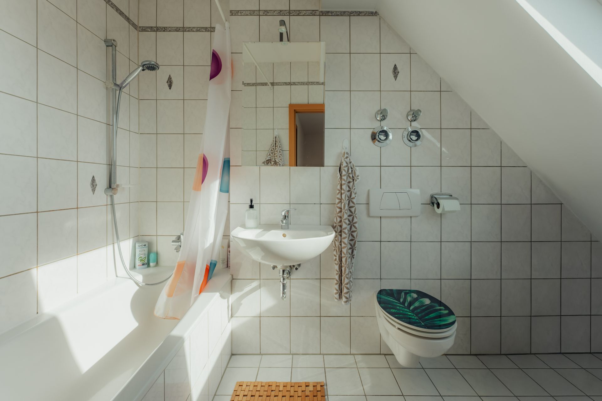 Helles Bad mit Badewanne, Duschvorhang, Waschbecken, Spiegel und Toilette unter einer Schräge.