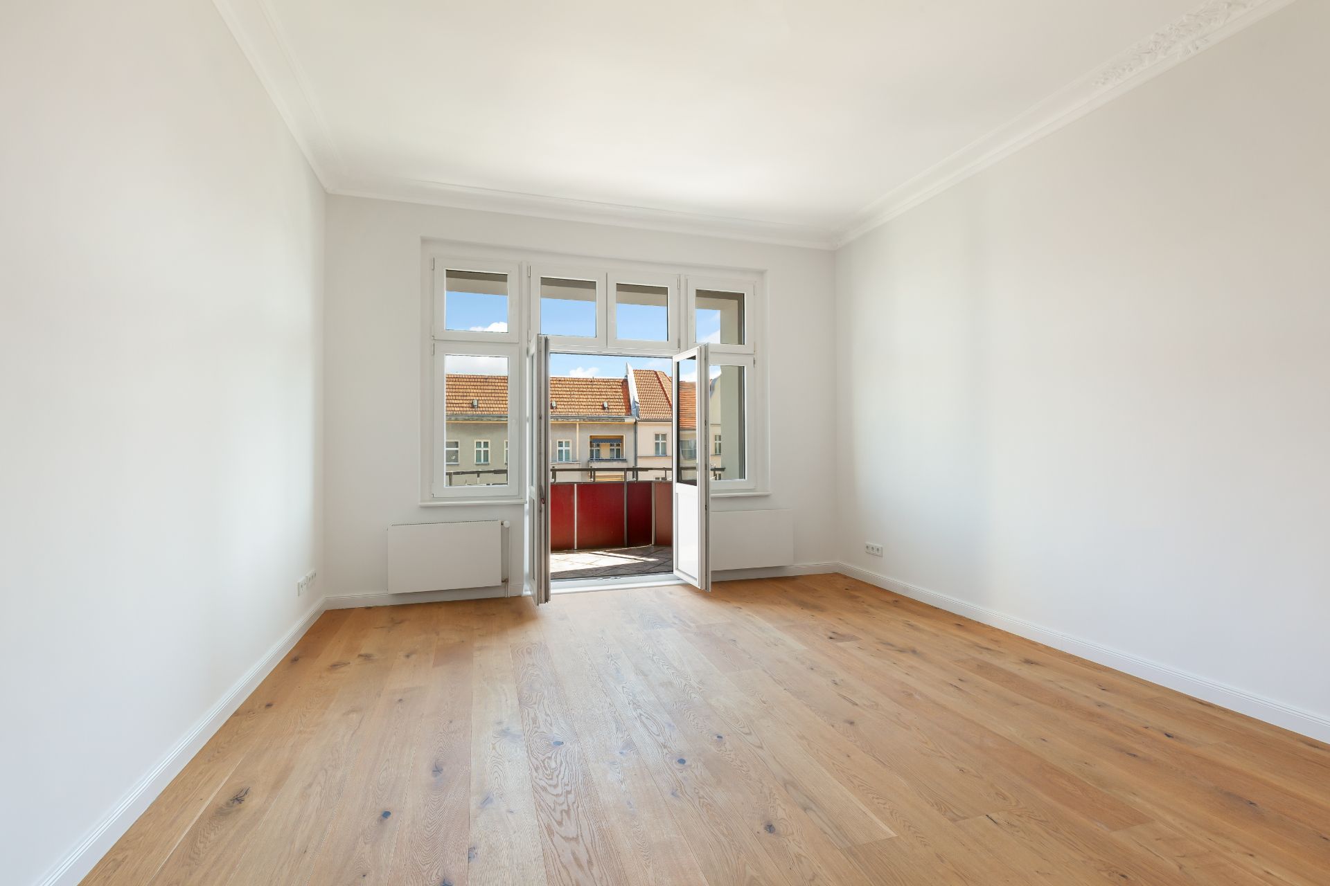 Heller, leerer Raum mit Holzfußboden und offenen Fenstertüren zu einem Balkon.