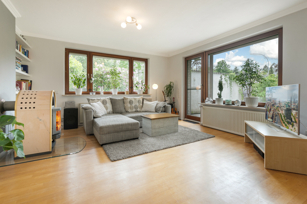 Helles Wohnzimmer mit Ecksofa, Holzofen, großen Fenstern und Pflanzen auf Fensterbänken.