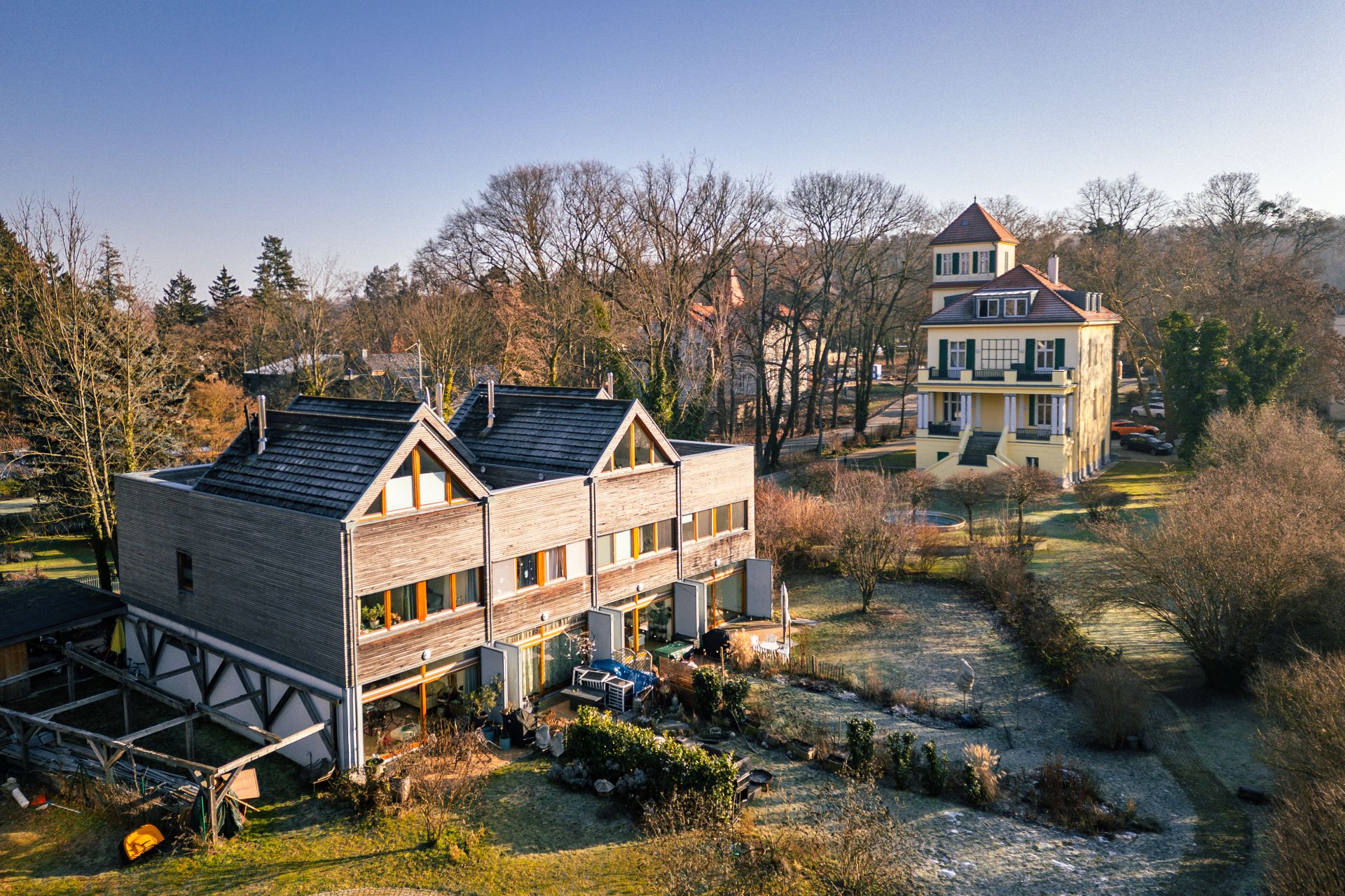 Zwei Wohnhäuser mit Gärten, im Vordergrund ein modernes Holzhaus, im Hintergrund ein gelbes Mehrfamilienhaus.
