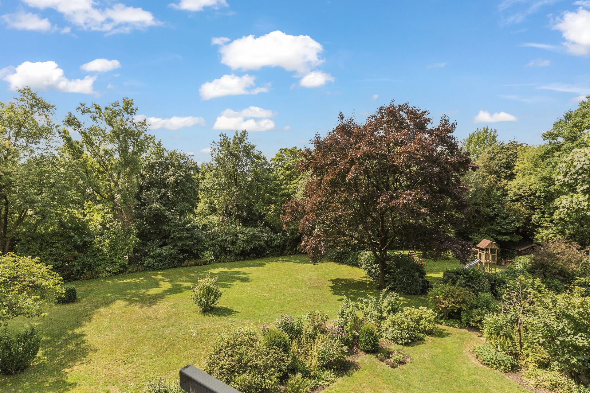 Großer Garten mit Rasen, Bäumen und Sträuchern, kleiner Holzspielplatz im Hintergrund.