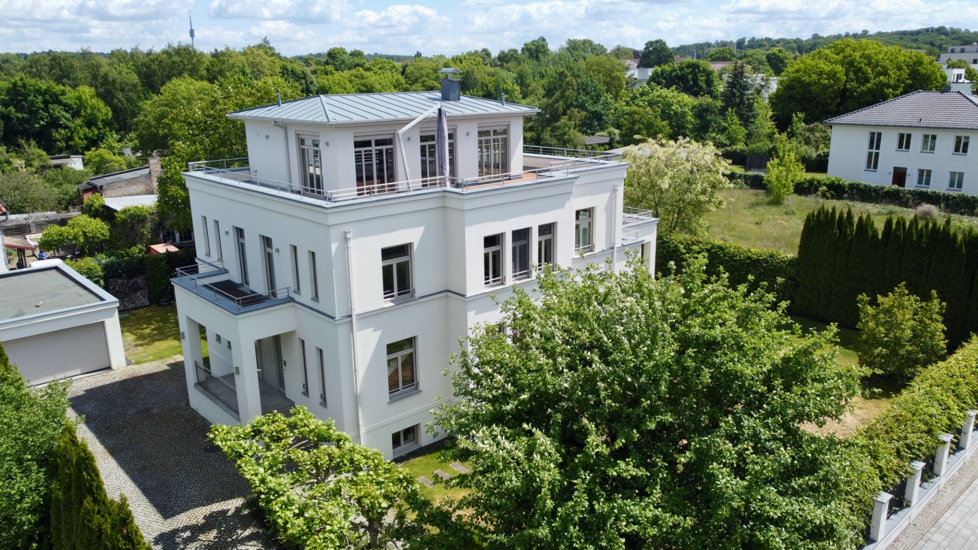 Modernes weißes Wohnhaus mit Flachdach, großem Balkon und umgeben von Bäumen und Garten.