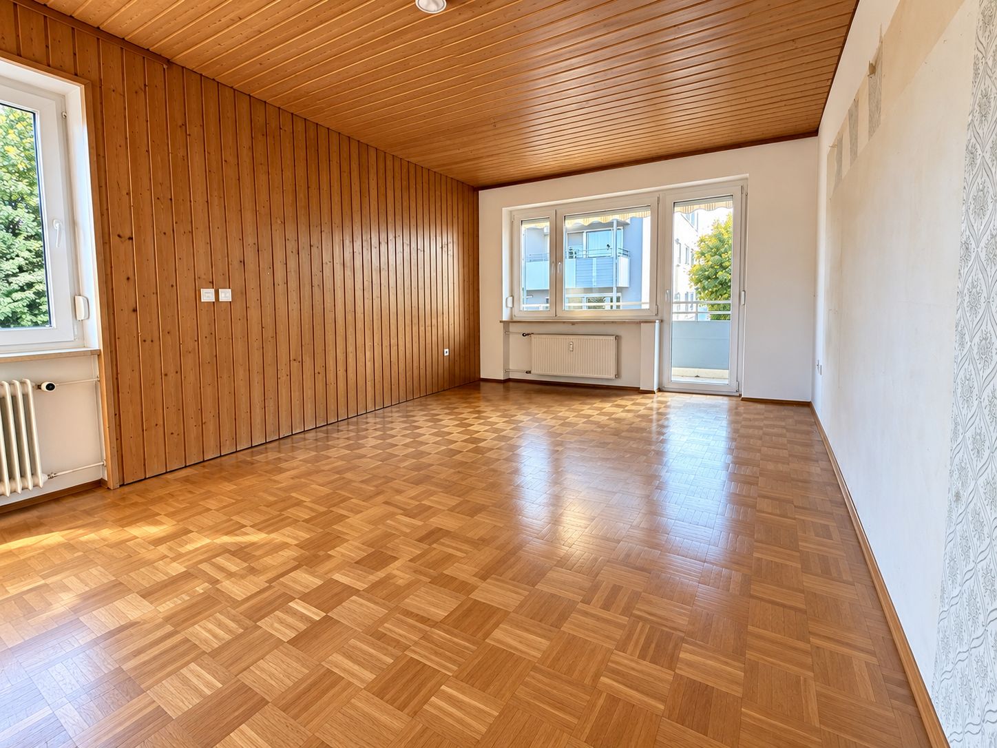 Helles Wohnzimmer mit Holzdecke, Holzwand, Parkettboden, Fenstern und Zugang zum Balkon.