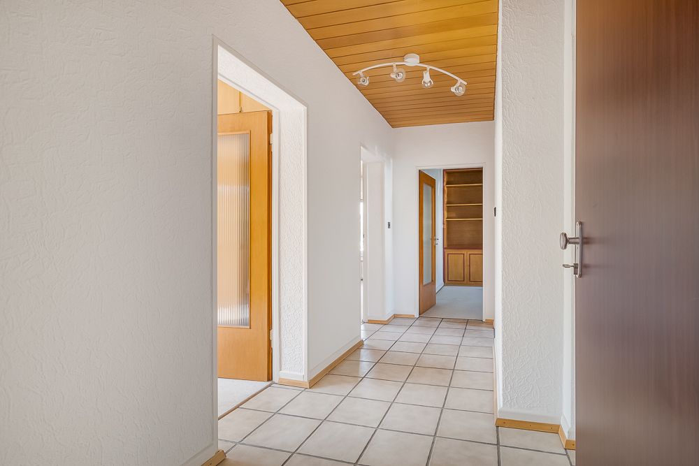 Flur mit Fliesenboden, weißen Wänden und Holztüren, Decke mit Holzverkleidung und Strahlerleuchte.