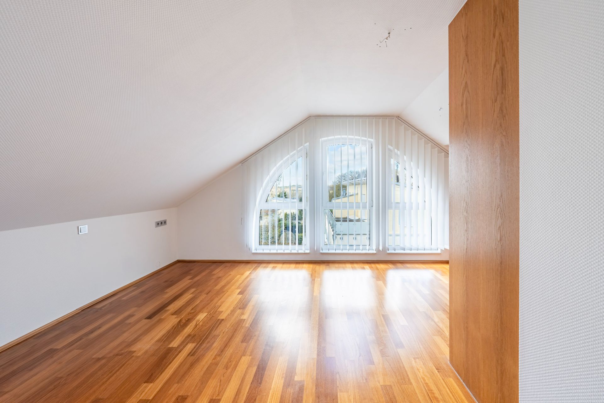 Heller Raum mit schräger Decke, großen Fenstern und Parkettboden.