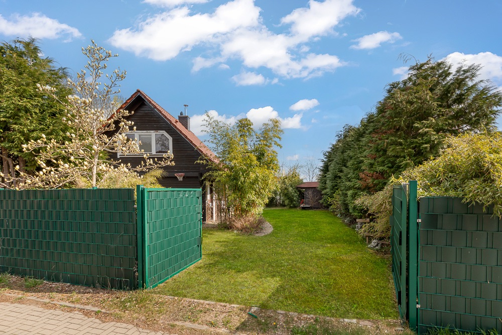 Eingezäunter Garten mit grünem Rasen, Bäumen und einem dunklen Haus mit Satteldach im Hintergrund.