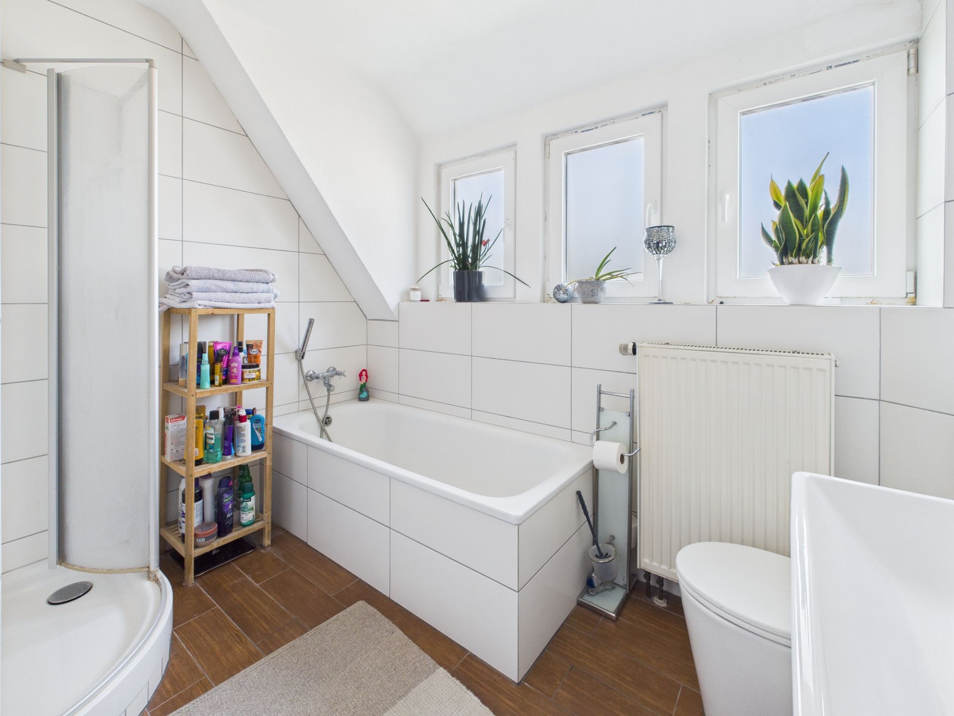 Helles Bad mit Badewanne, Dusche, Regal, WC und drei Fenstern mit Pflanzen.