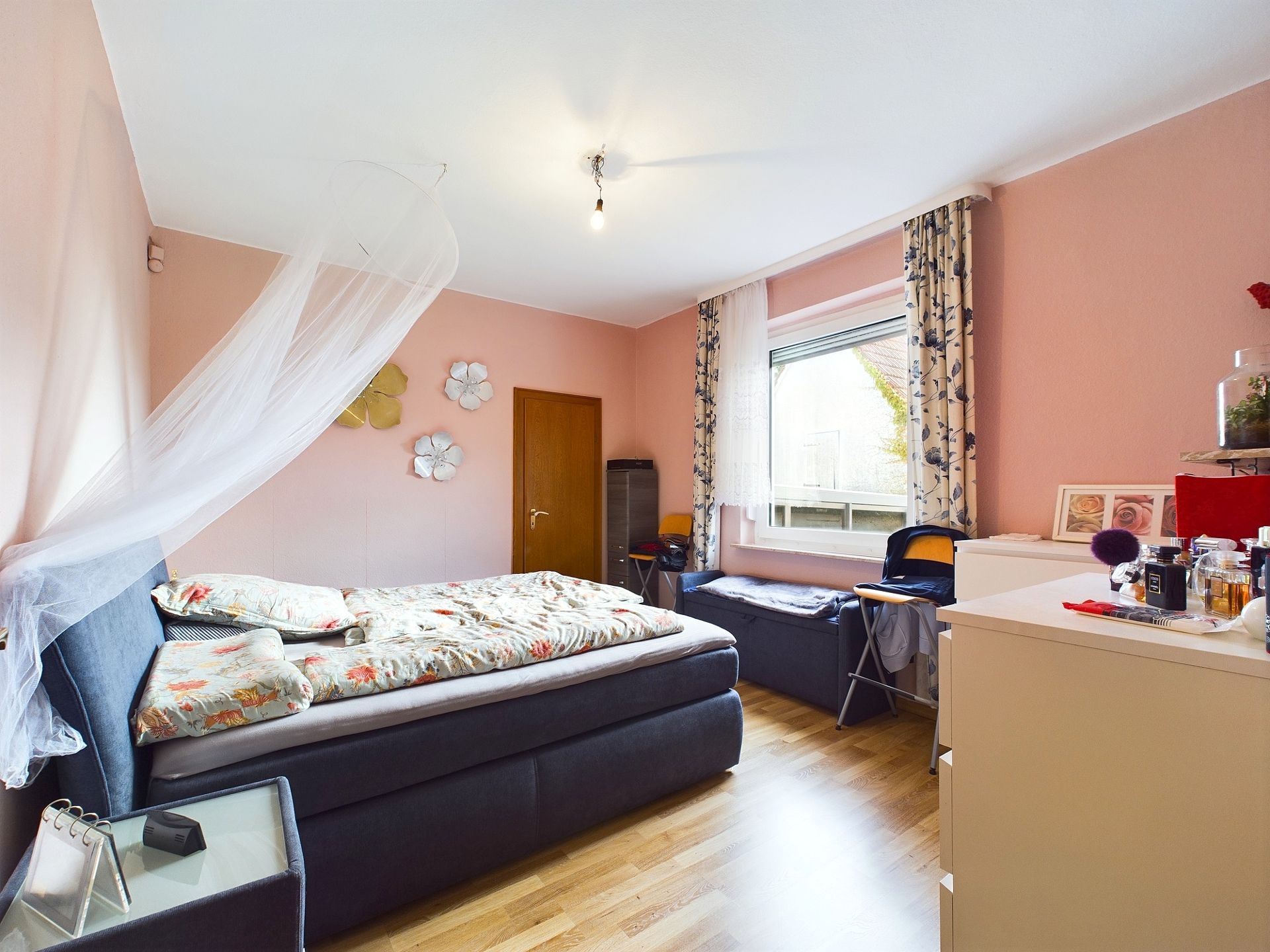 Helles Schlafzimmer mit Doppelbett, rosa Wänden, Fenster, Kommode und Dekorationen an der Wand.