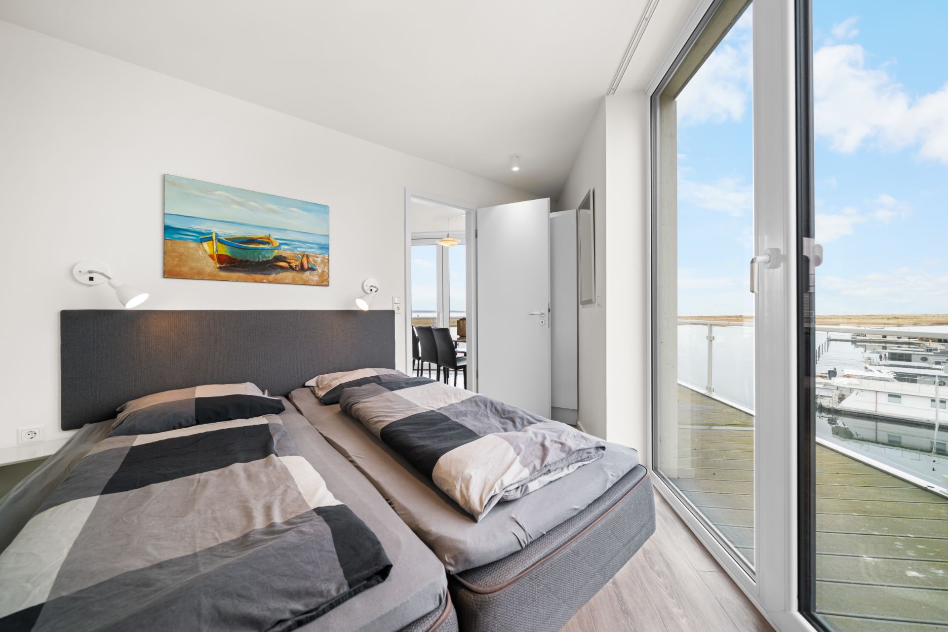 Helles Schlafzimmer mit zwei Einzelbetten, großem Fenster und Zugang zu einem Balkon mit Blick auf einen Hafen.