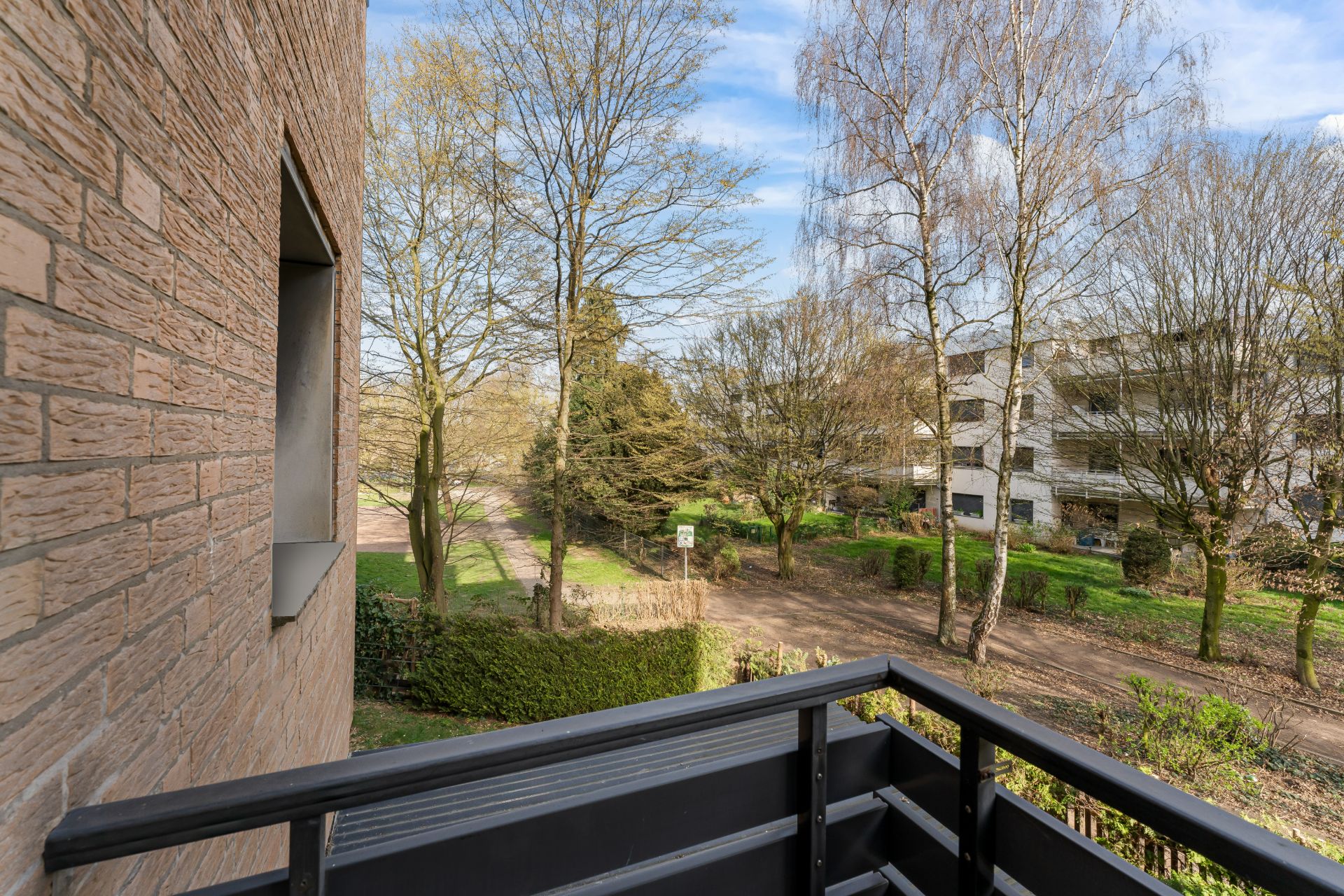 Blick von einem Balkon auf Bäume und einen Weg, daneben ein Wohngebäude.