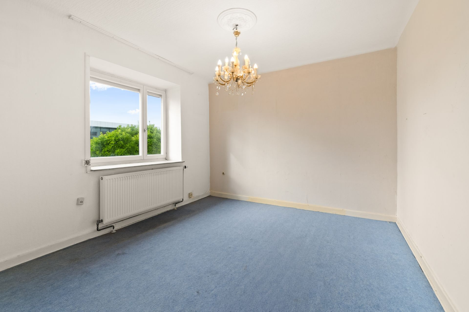Leerer Raum mit blauem Teppich, Fenster, Heizkörper und Kronleuchter an der Decke.