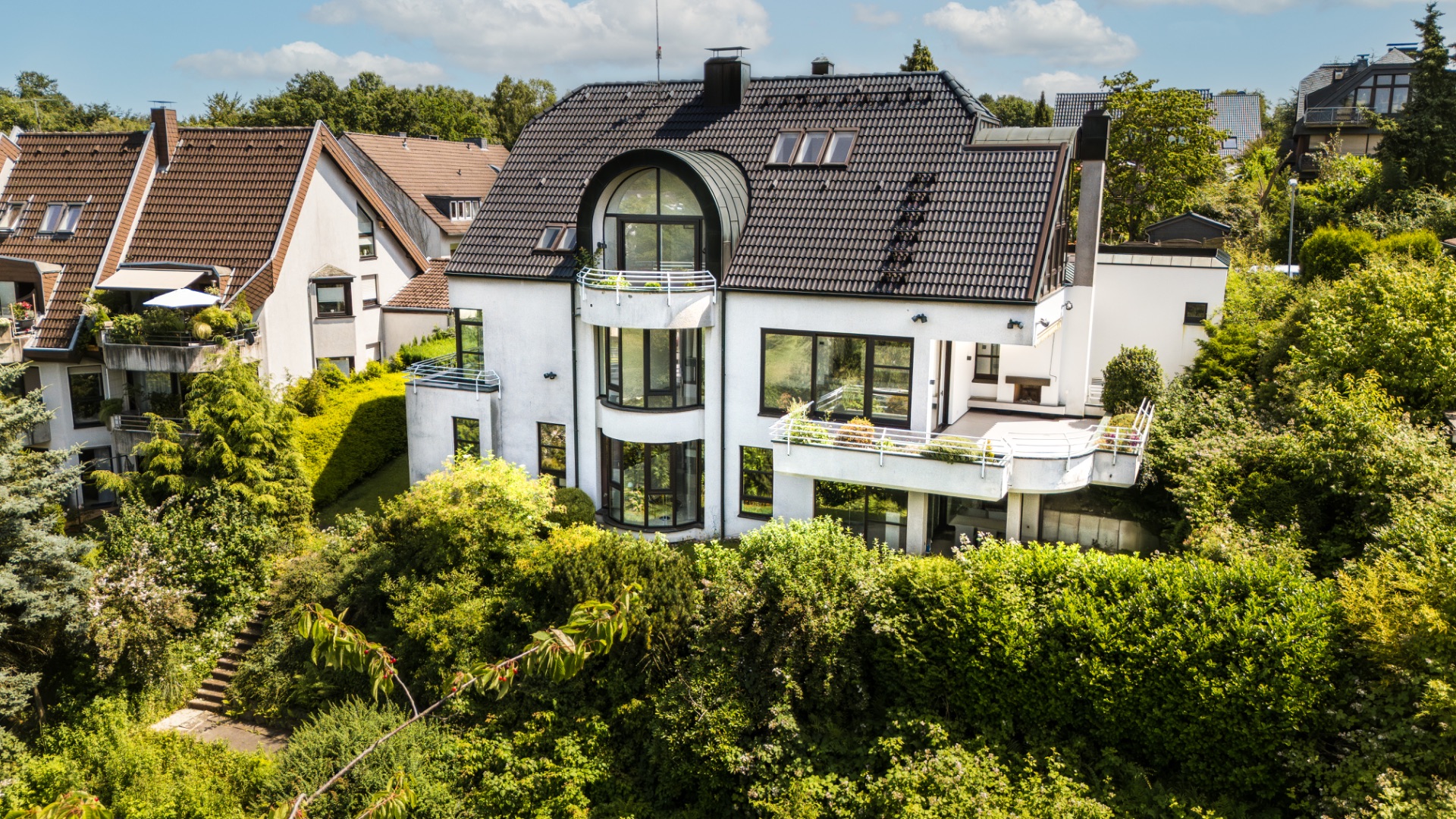 Modernes weißes Wohnhaus mit Balkon und großen Fenstern, umgeben von viel Grün.